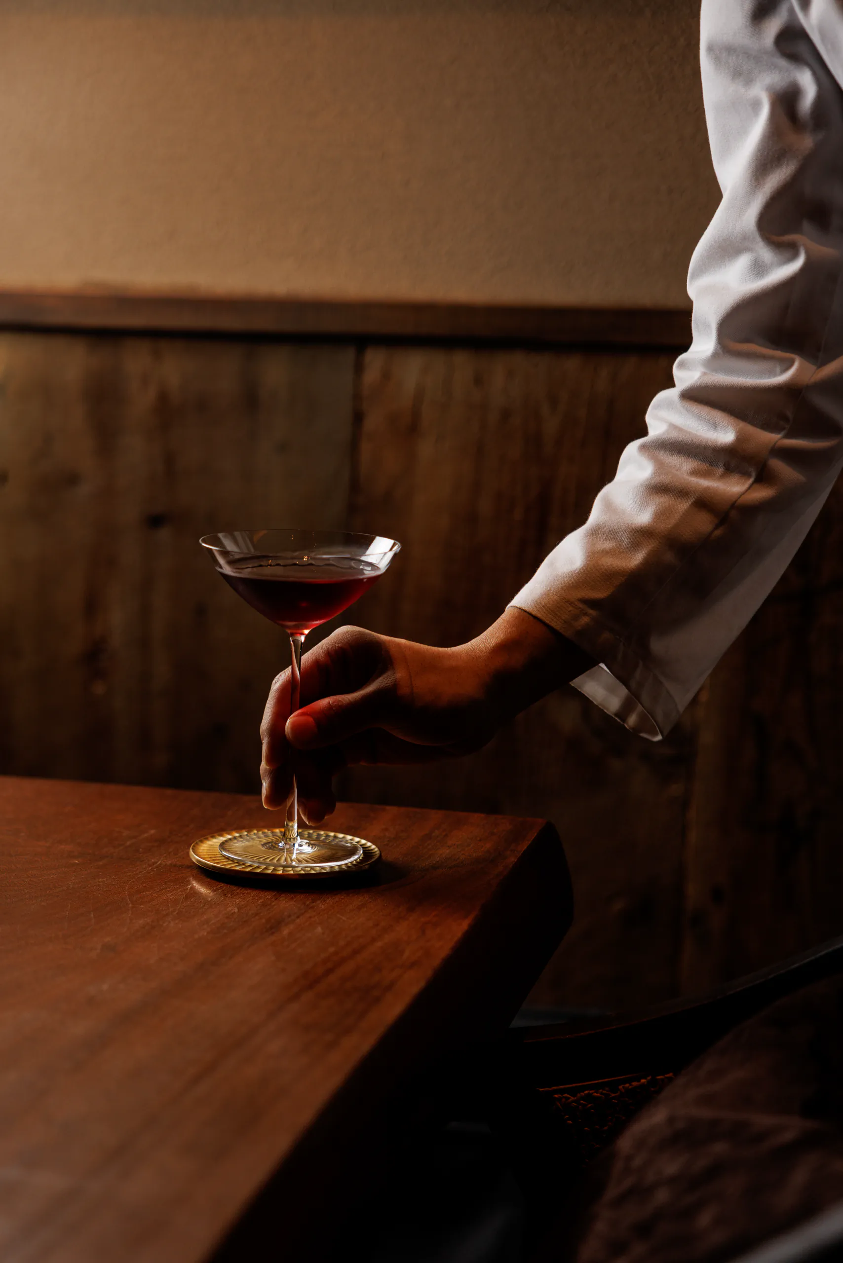 Hand setting down a dark cocktail on a wooden bar in dim light in a small cocktail bar in Tokyo