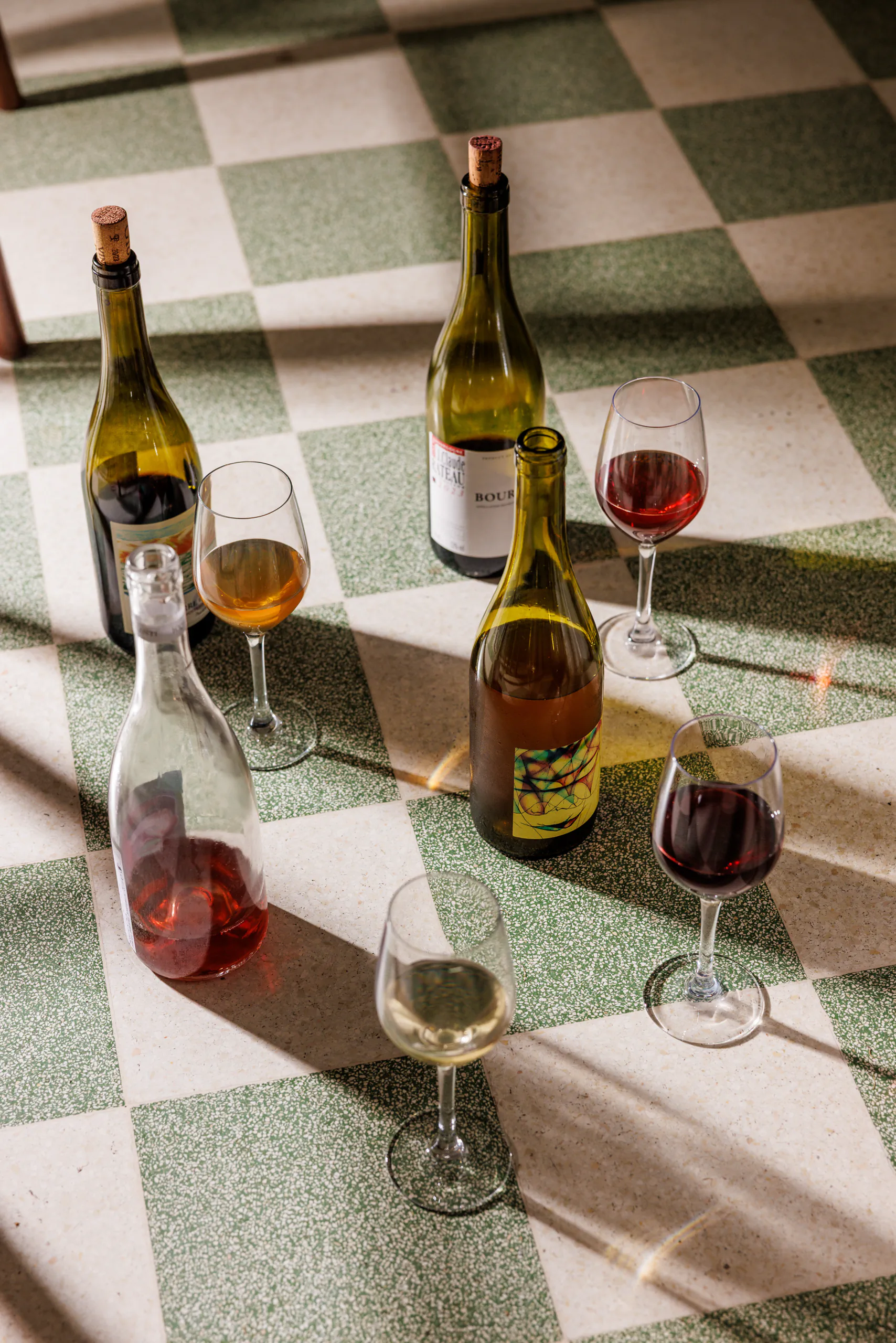Open bottles of white and red wine with glasses on a sunlit checkerboard floor