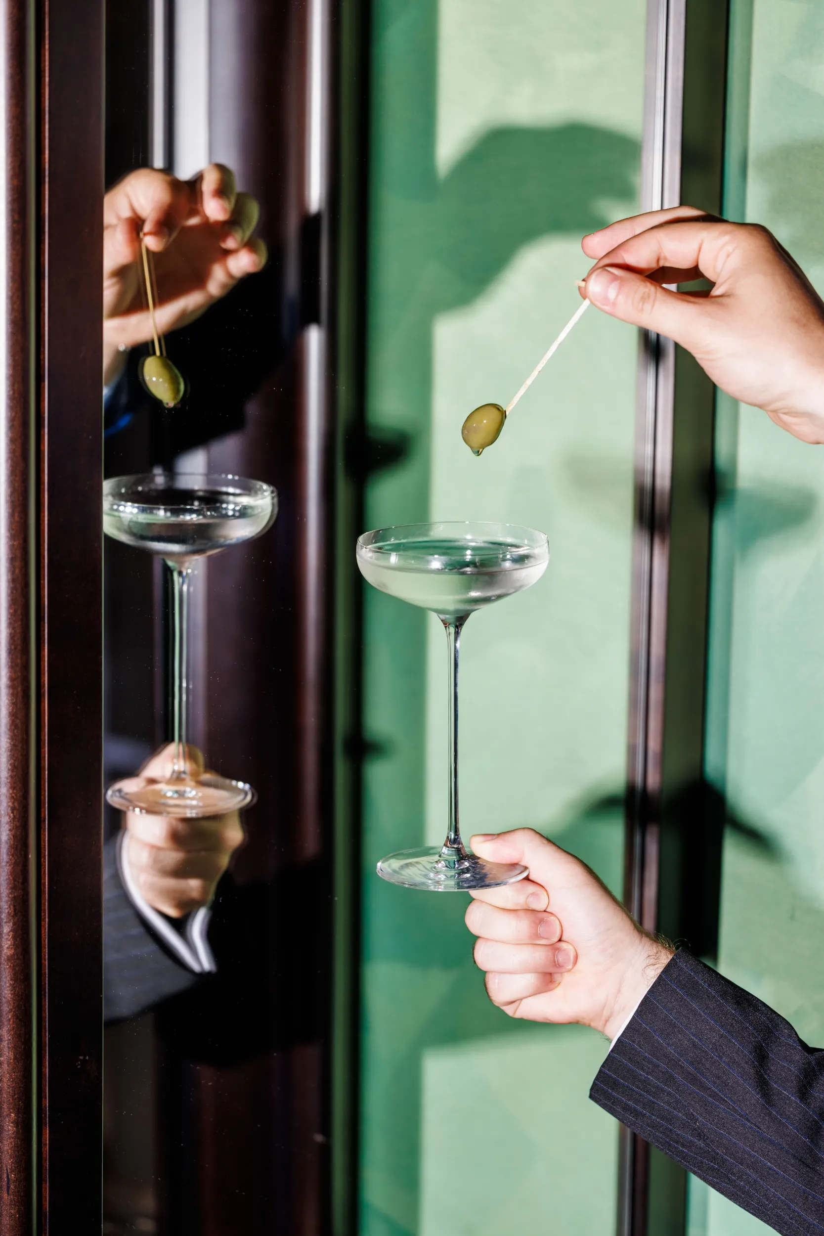 Two hands garnishing coupe cocktails with olives, reflected against a green surface at Grand Hotel Flora, Rome, Italy