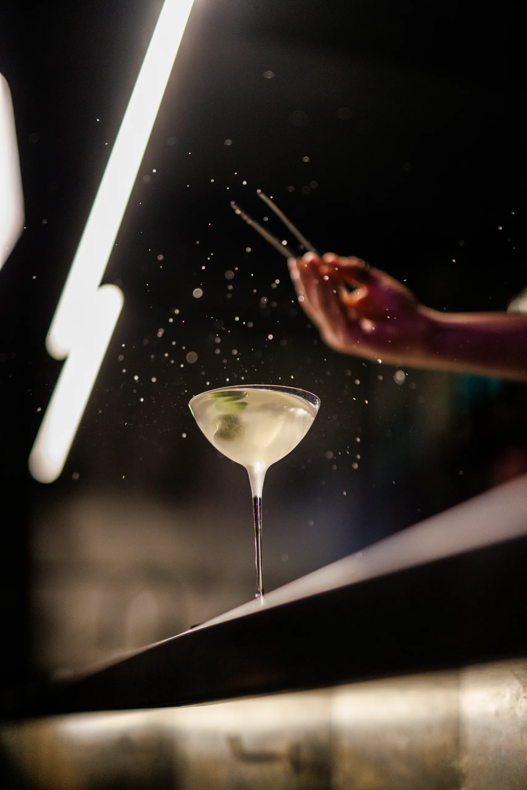 Martini on a bar being garnished with tweezers amid streaks of light