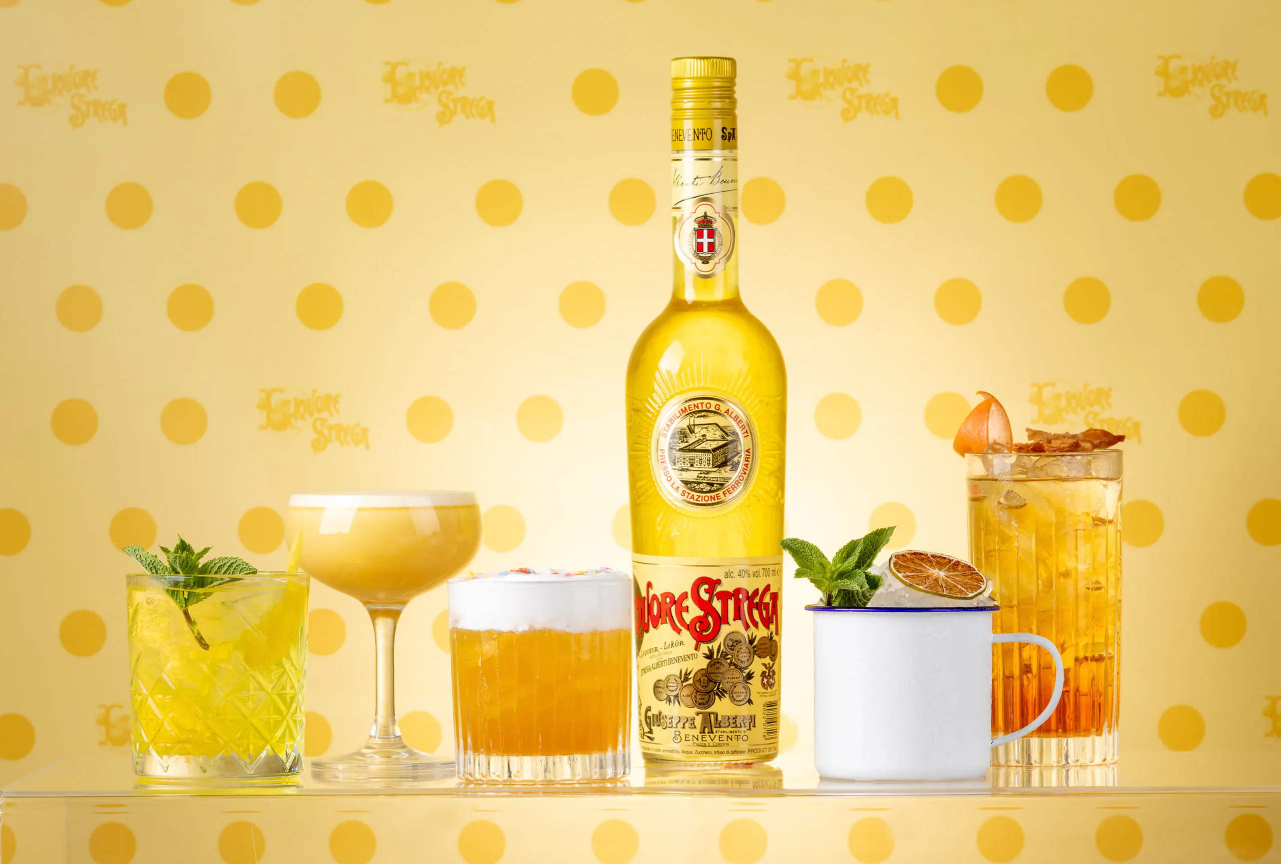Still life of cocktails around a Liquore Strega bottle on a yellow polka-dot background