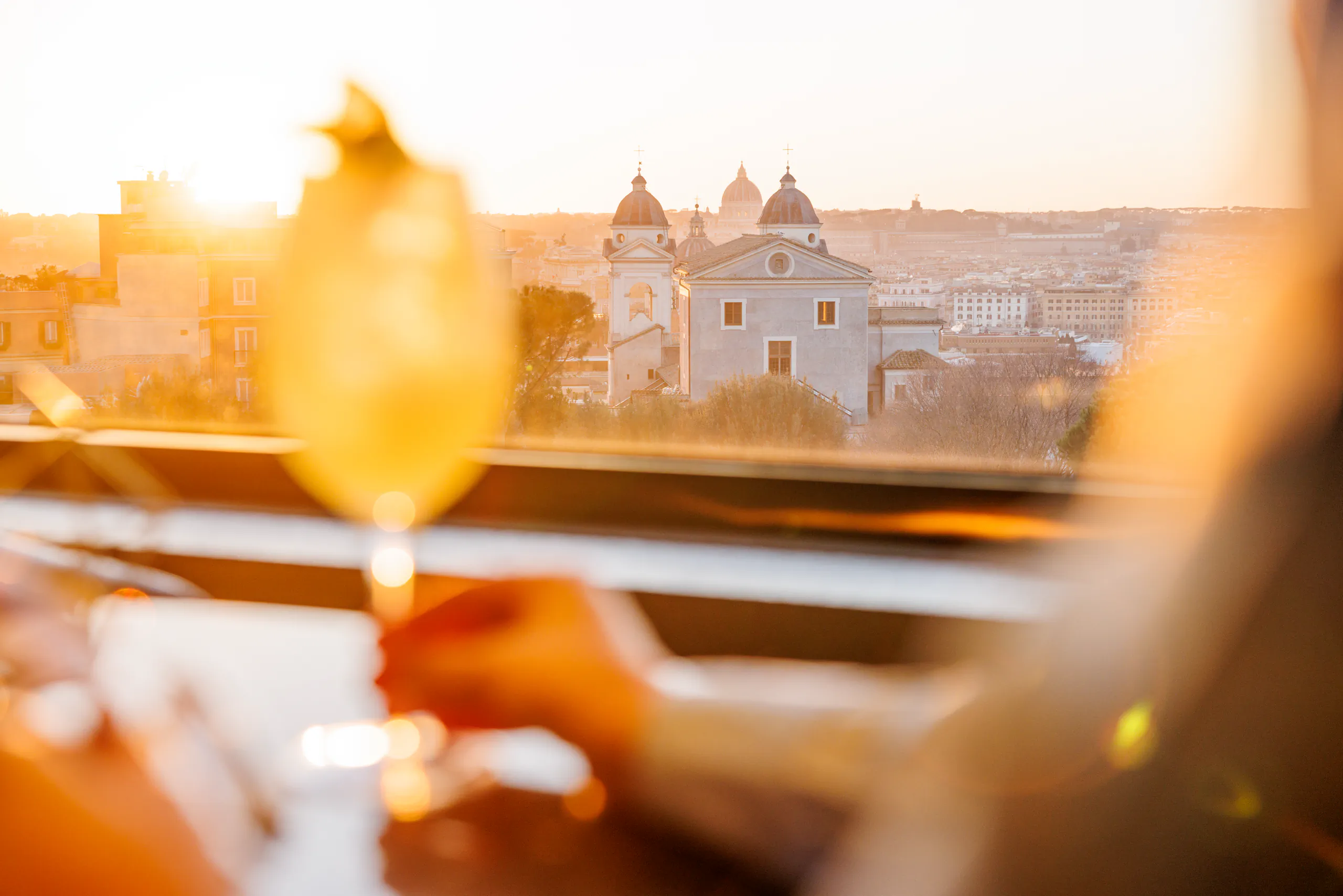 Hand holding an orange cocktail on a rooftop terrace at sunset over a city