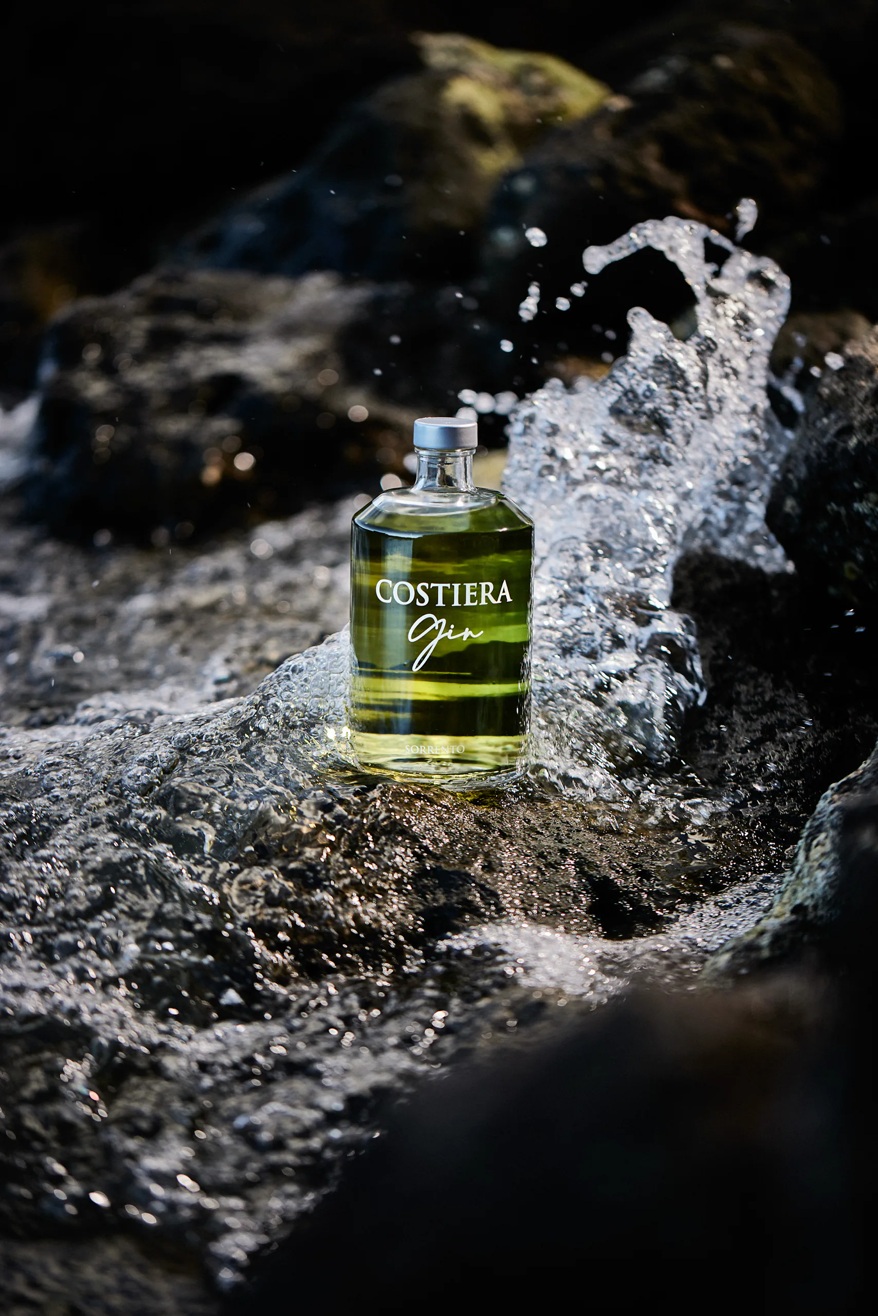 Bottle of Costiera Gin on wet black rocks amid splashing water — Gin Costiera