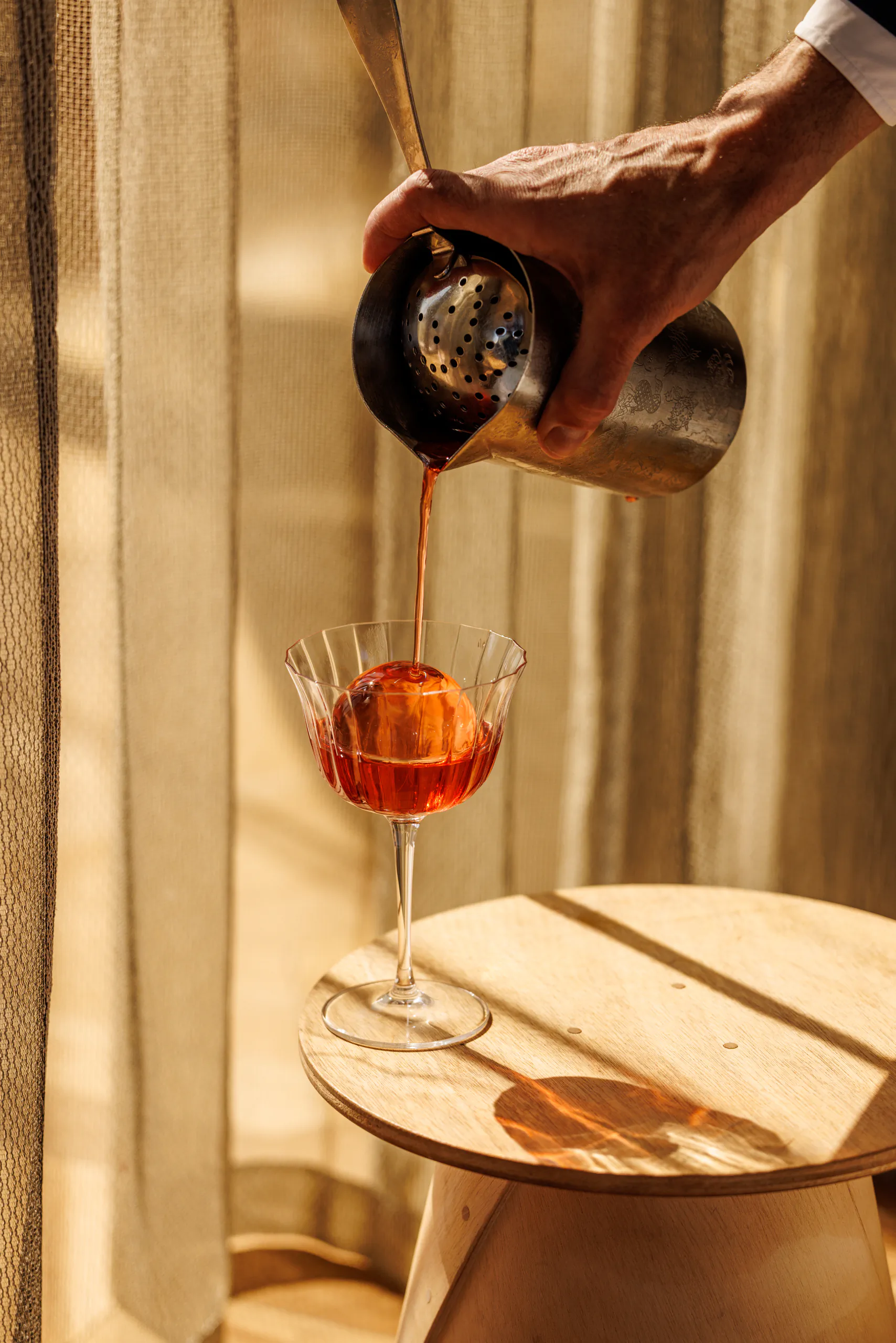 Hand pouring a red cocktail into a coupe on a small stool by a sunlit curtain — Horto, Milan