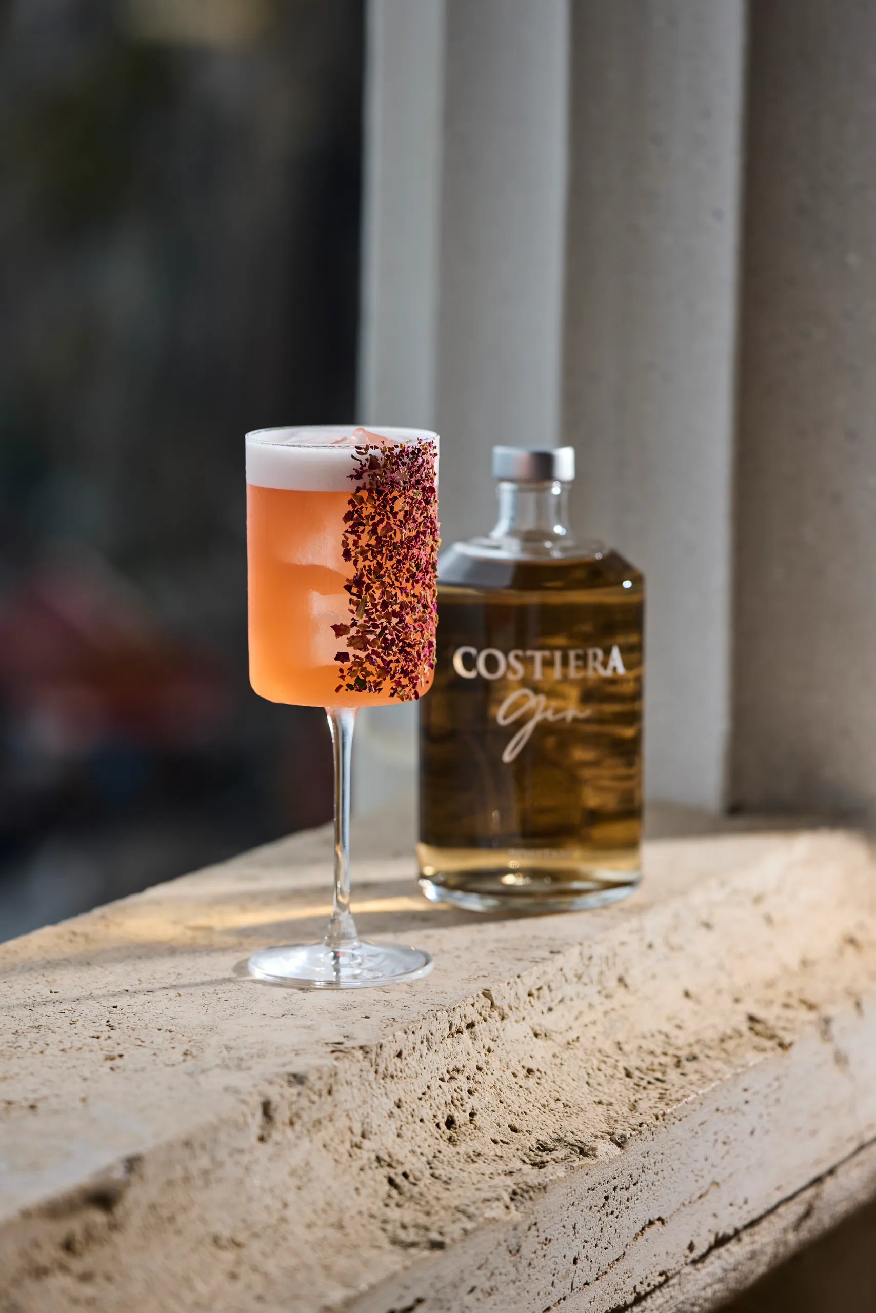 Pink cocktail with a spiced rim beside a Costiera Gin bottle on a stone ledge — Bellevue Syrene, Sorrento