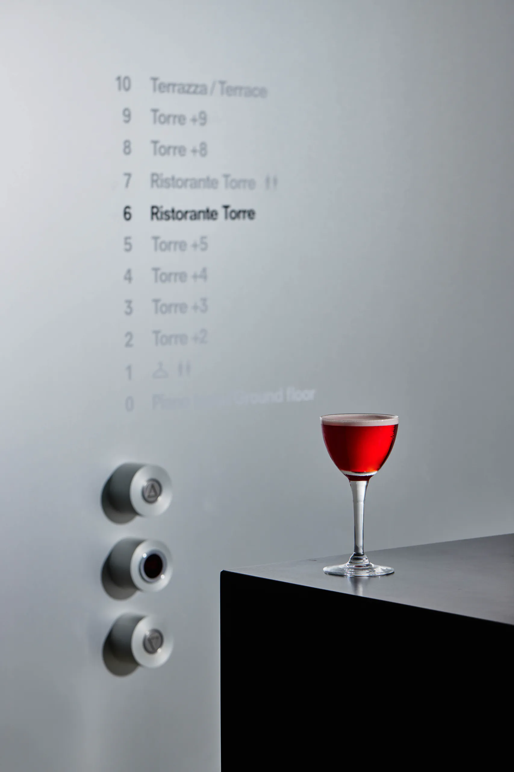 Red cocktail on a ledge beside an elevator floor directory panel — Torre, Fondazione Prada, Milan