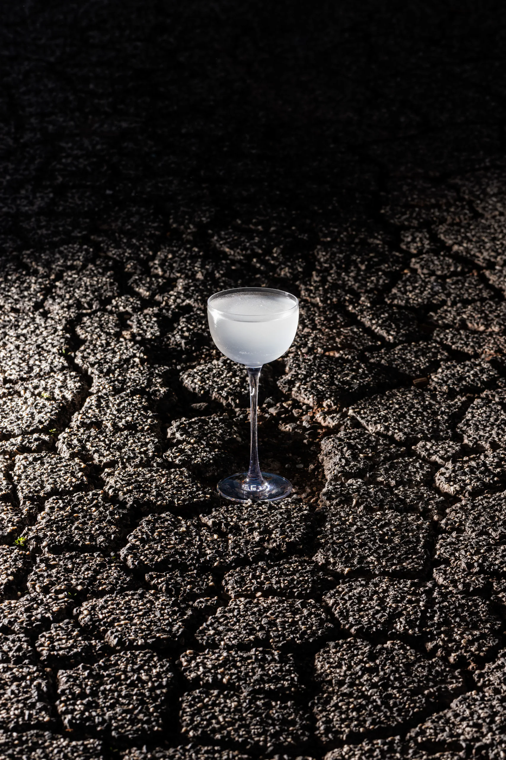 Pale cocktail in a coupe glass set on cracked dark asphalt