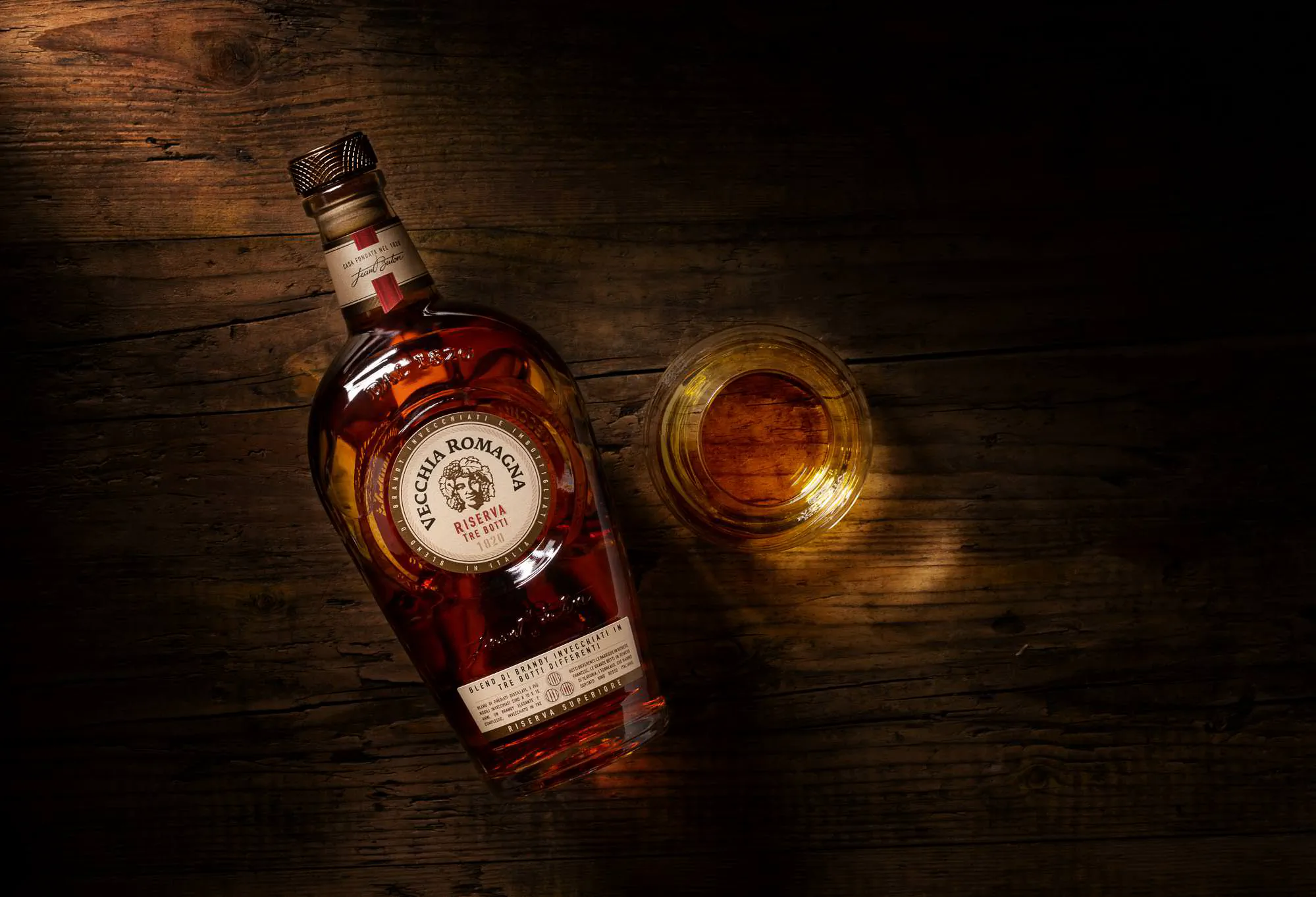 Bottle of Vecchia Romagna brandy on its side beside a glass of the spirit on dark wood