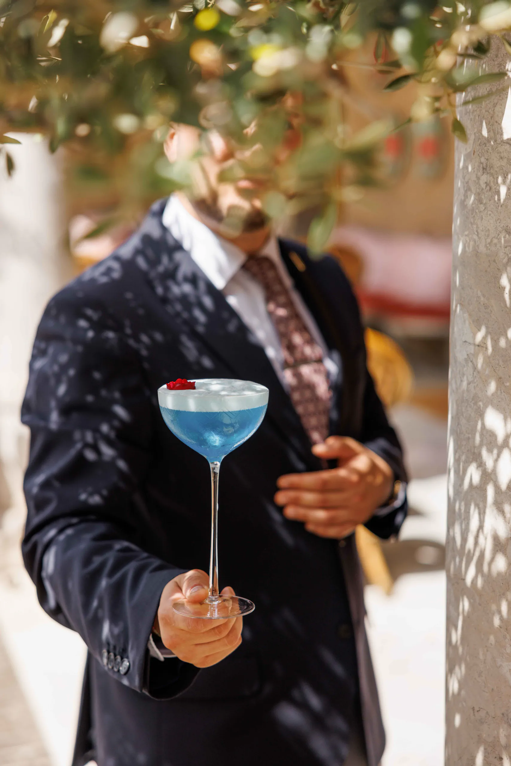 Man in a dark suit holding out a blue cocktail in a coupe glass by an olive tree at Villa Igiea a Roccoforte Hotel in Palermo, Italy