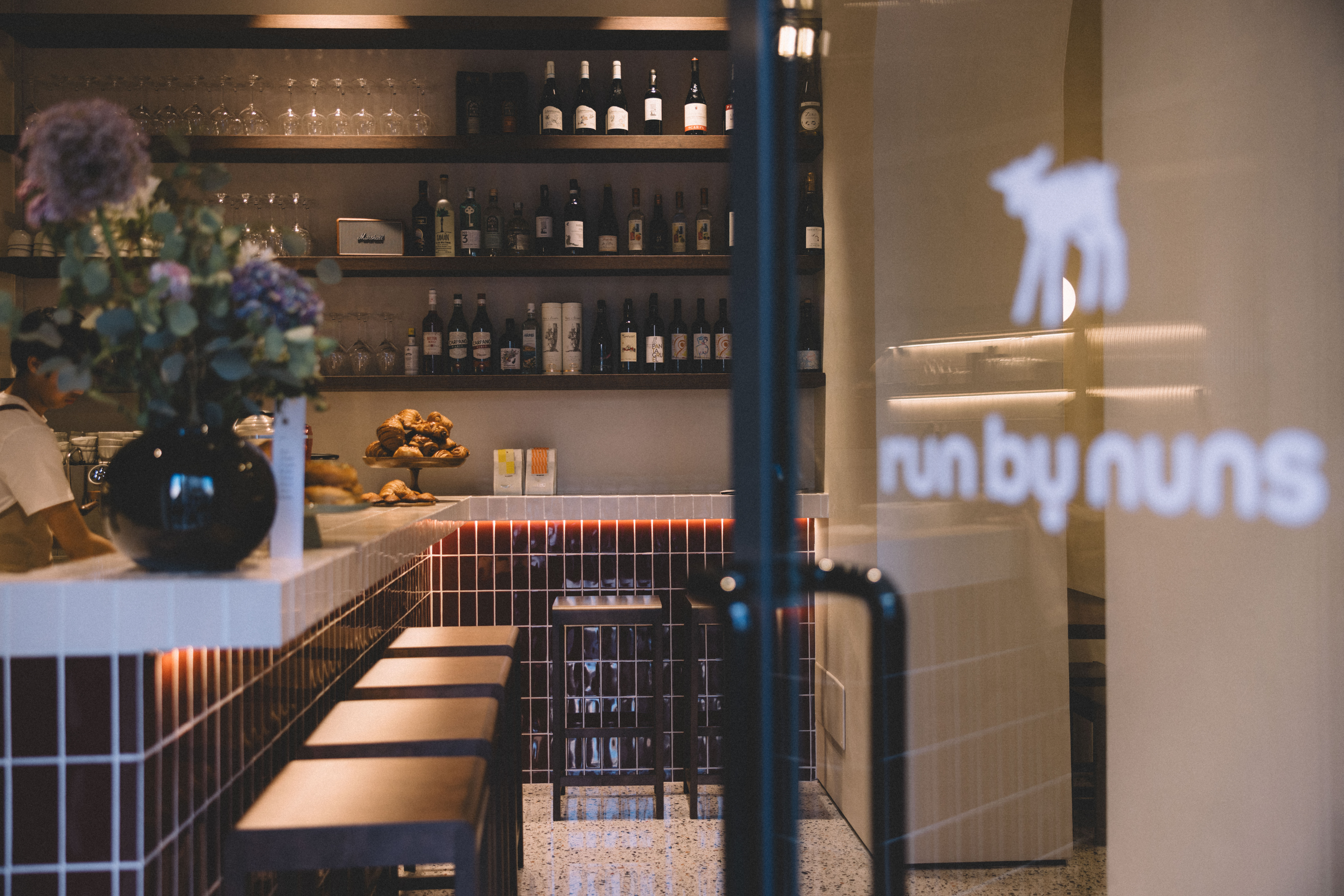 Bar interior with shelves of bottles, a tiled counter and a neon sign reading 'run by nuns'