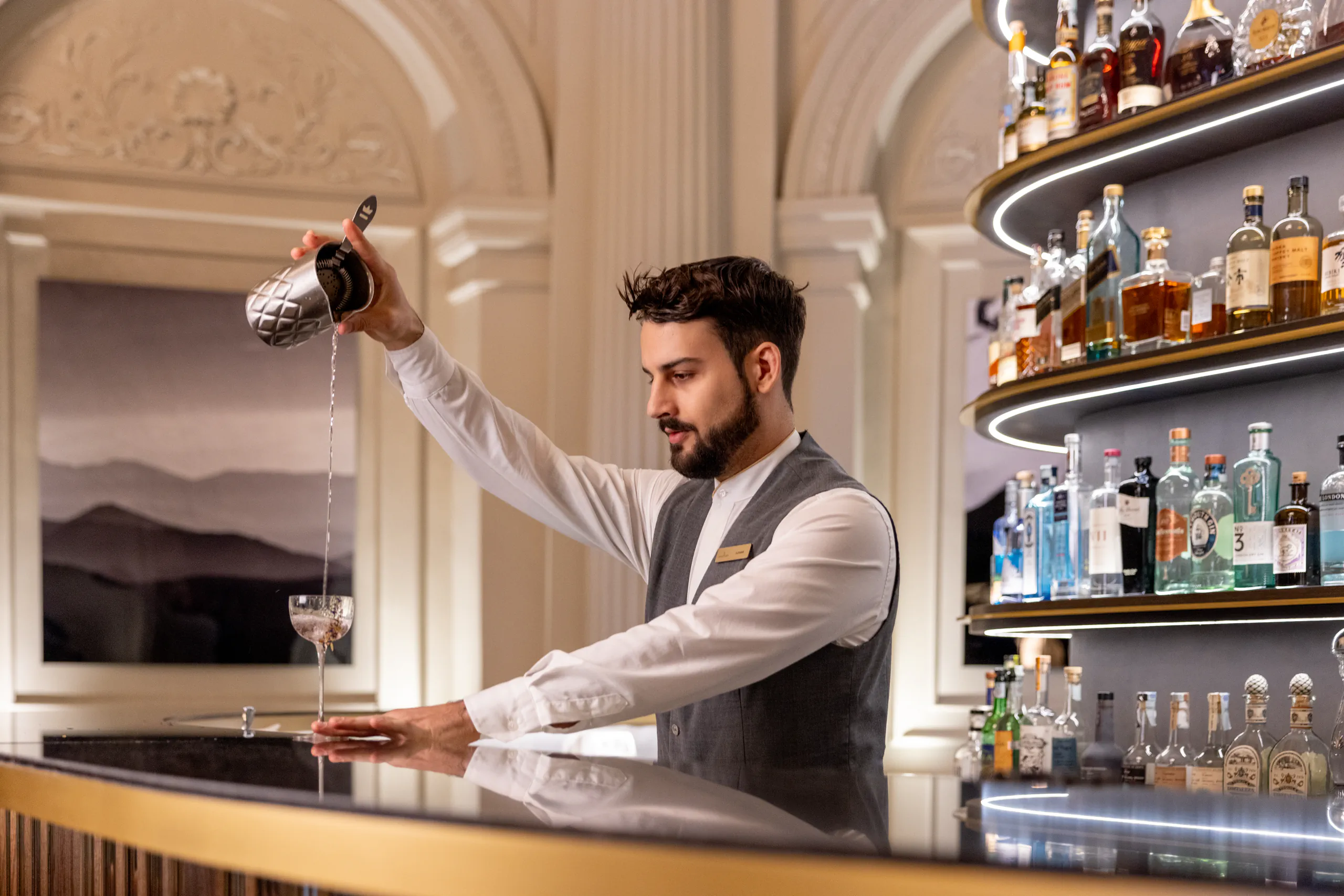 Bartender in a waistcoat pouring a drink behind an elegant bar lined with spirits