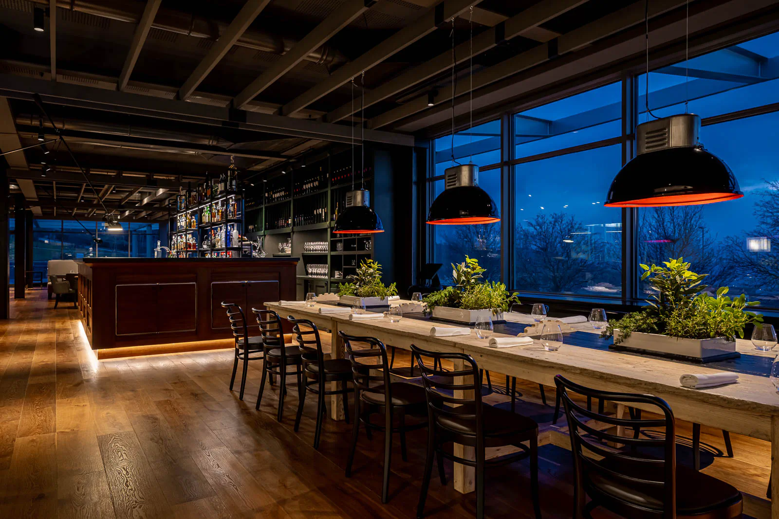 Restaurant with a long communal table and pendant lights overlooking the city at dusk