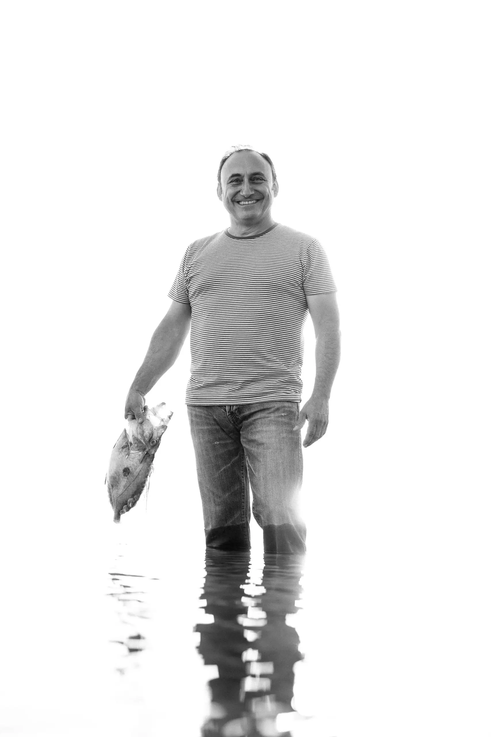 Black-and-white photo of a man walking in shallow water holding a fish