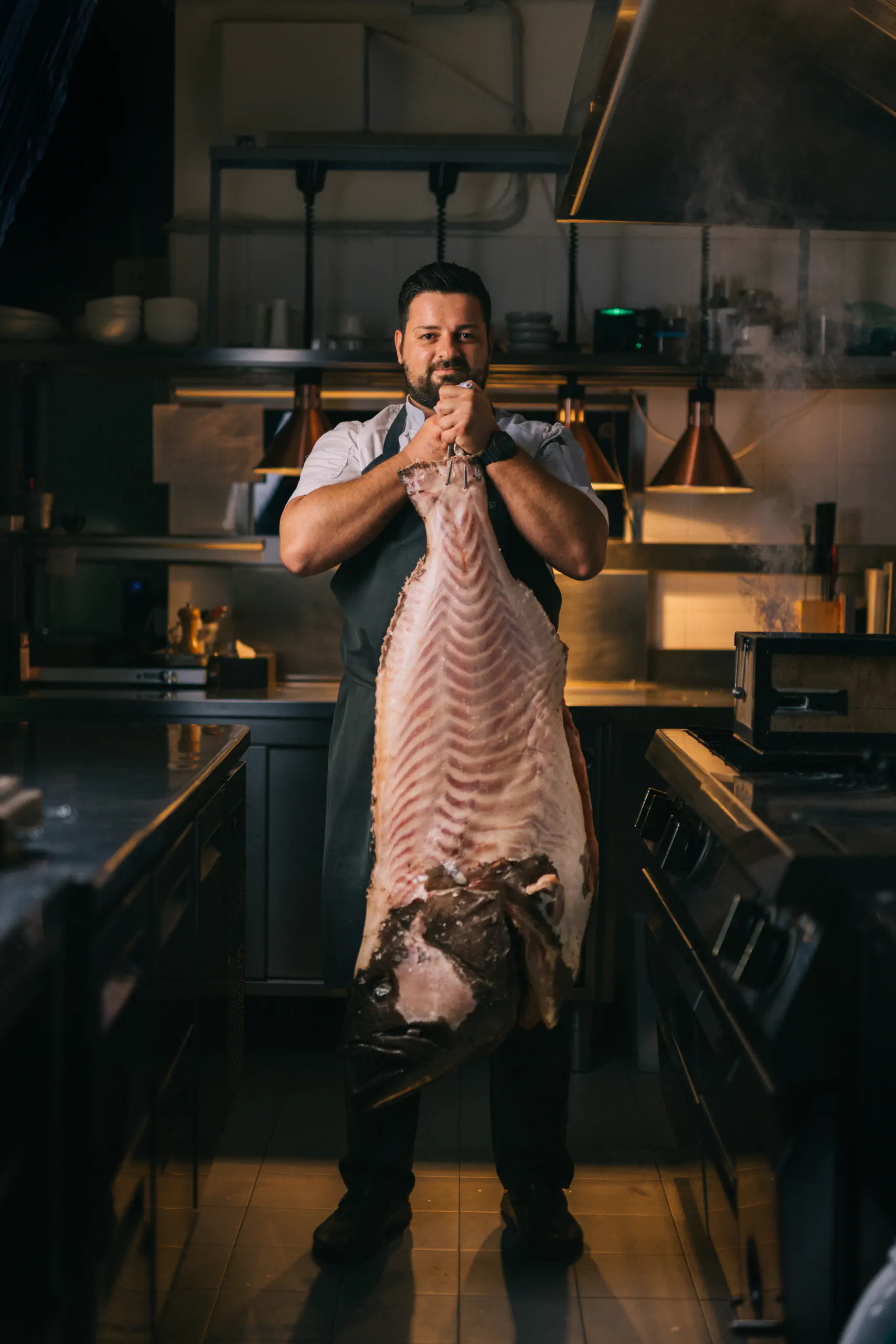 Man in an apron holding a large fish in a dark kitchen