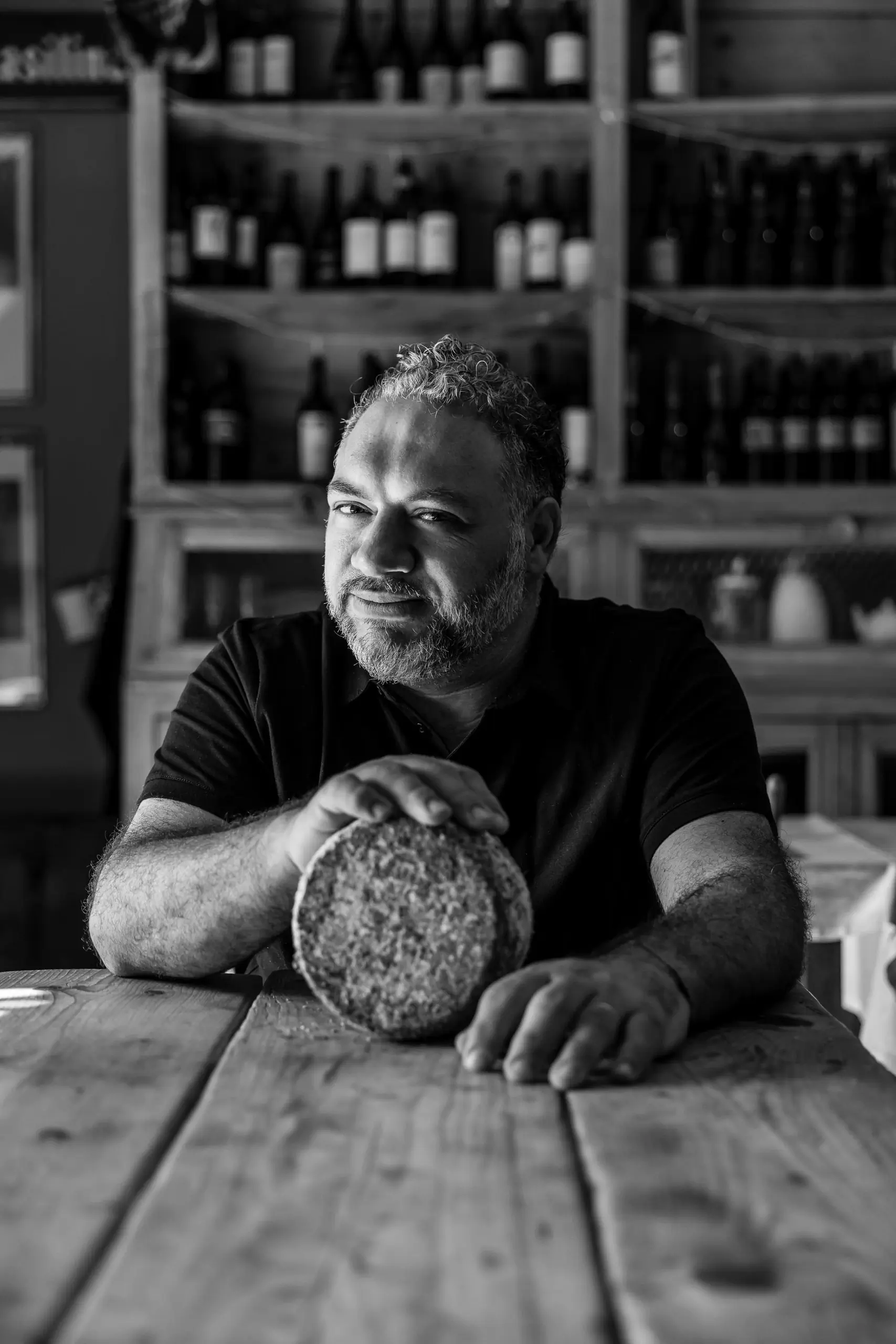 Black-and-white photo of a man holding a wheel of cheese at a table