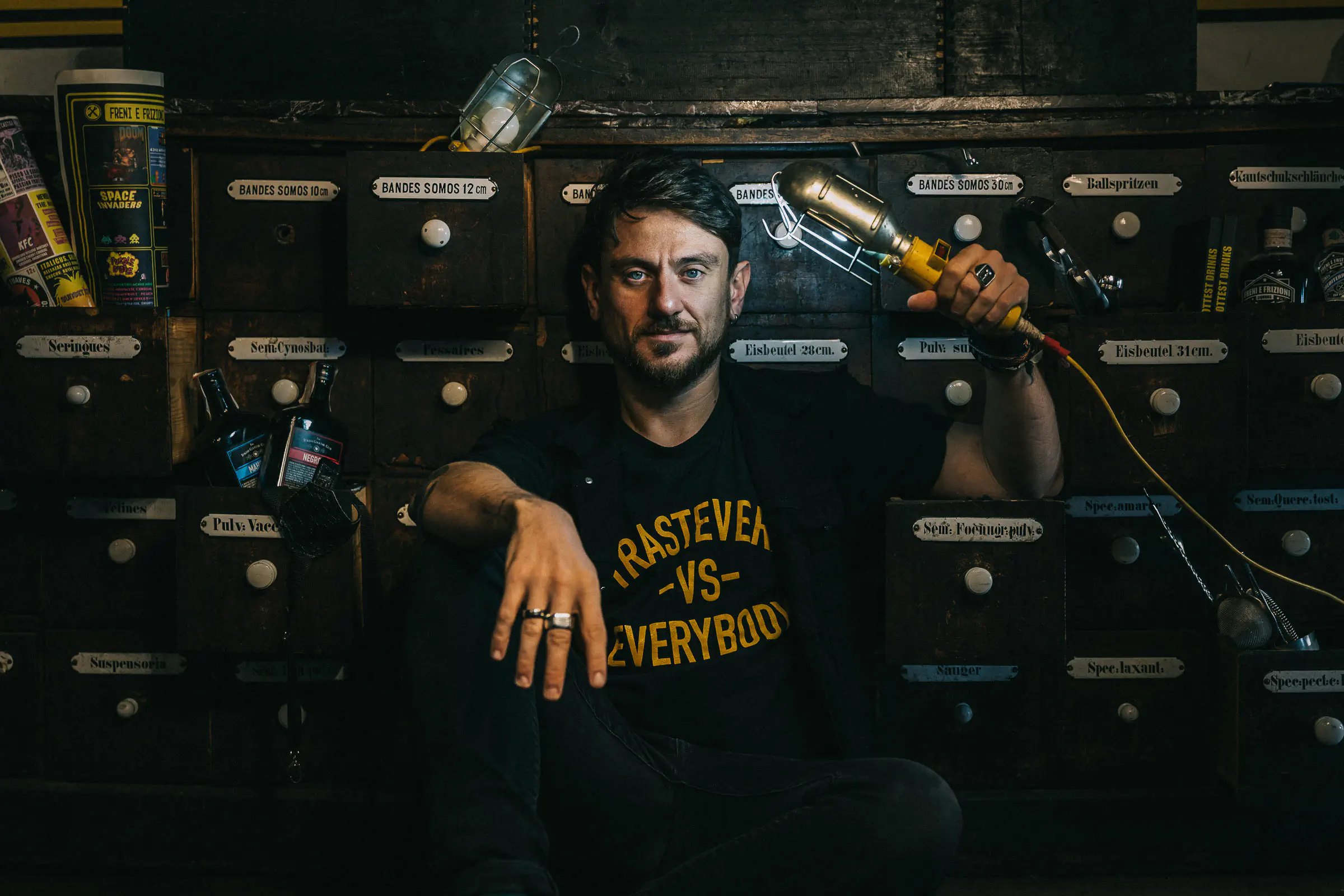 Man in a printed t-shirt seated in a bar with wooden drawers behind