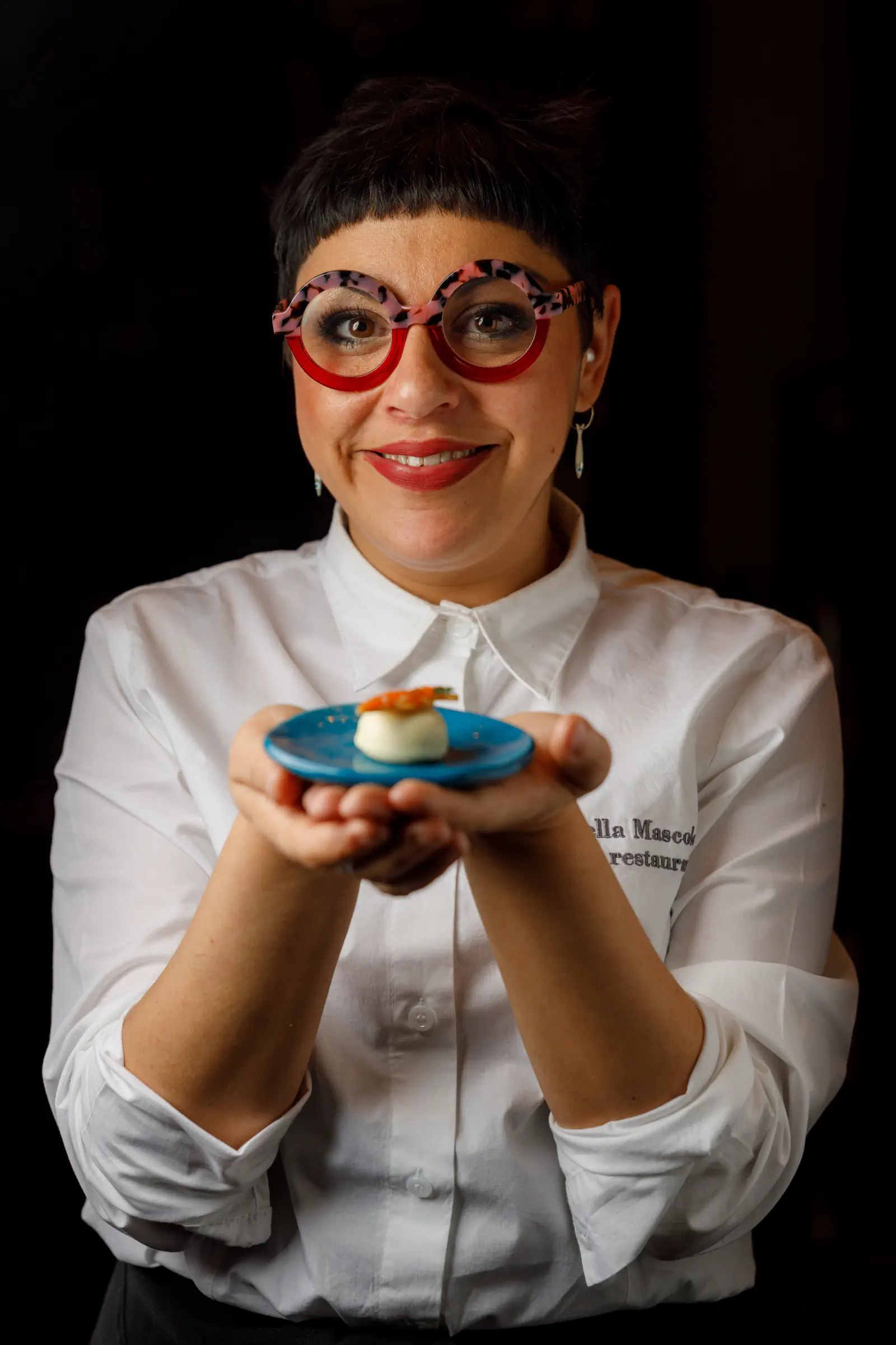 Woman in whites with red glasses holding out a small dish