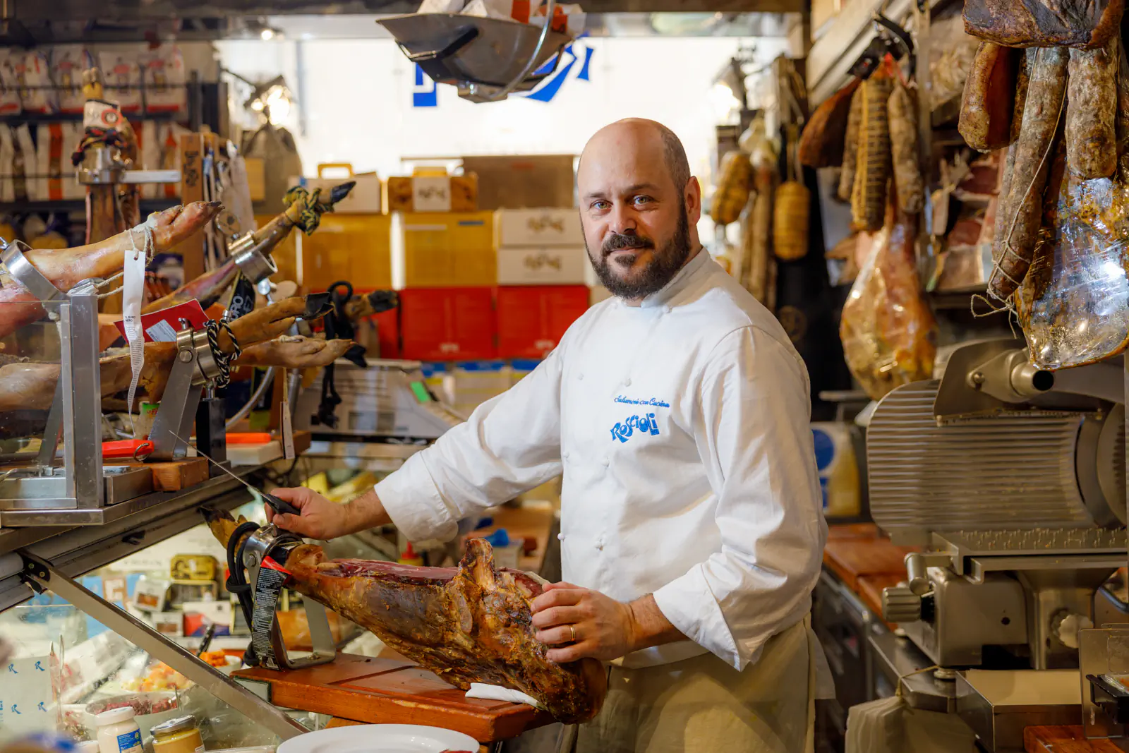 Man in whites at a deli counter with cured hams hanging behind