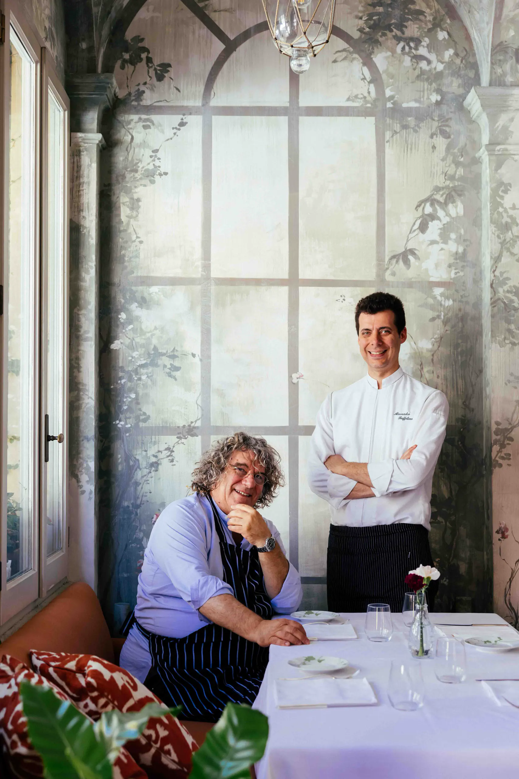 Woman seated and a chef standing at a table in a grand room with tall windows