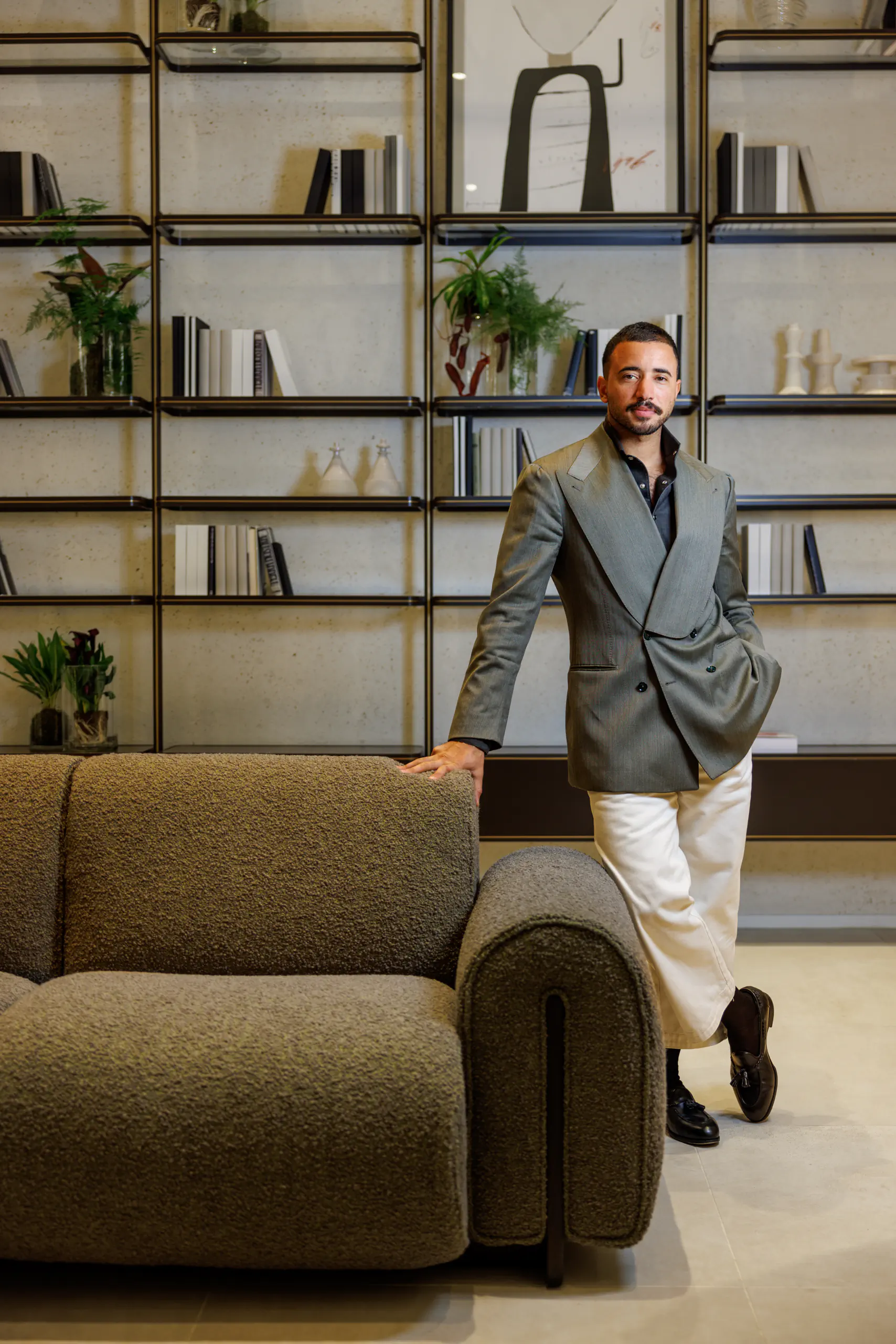 Man in a blazer standing by an armchair in front of a bookshelf