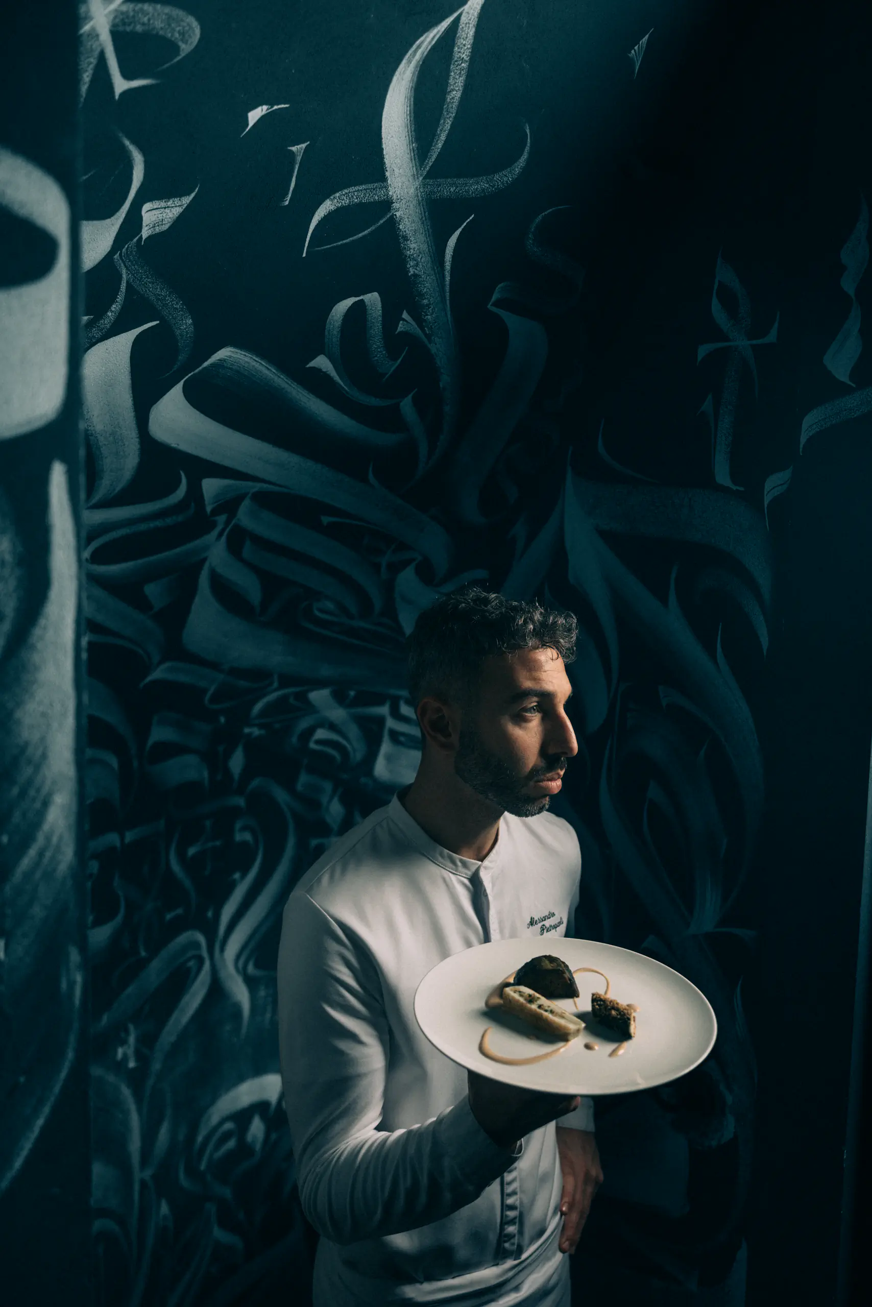 Man in whites holding a plate against a dark decorative backdrop