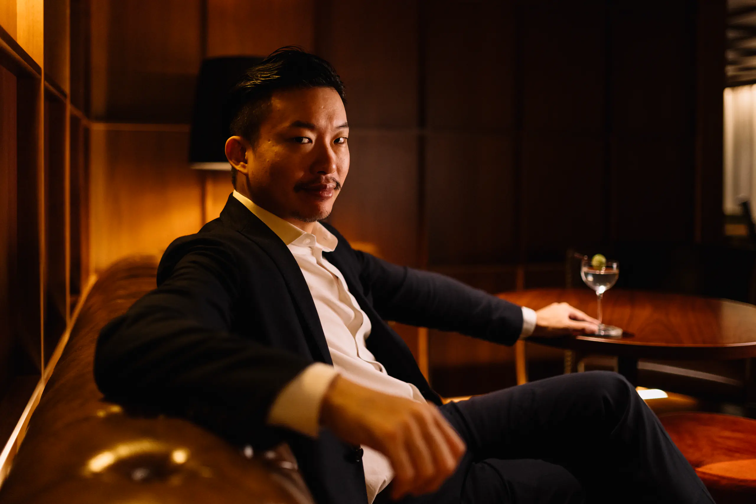 Man in a blazer seated in a warm-lit lounge holding a drink
