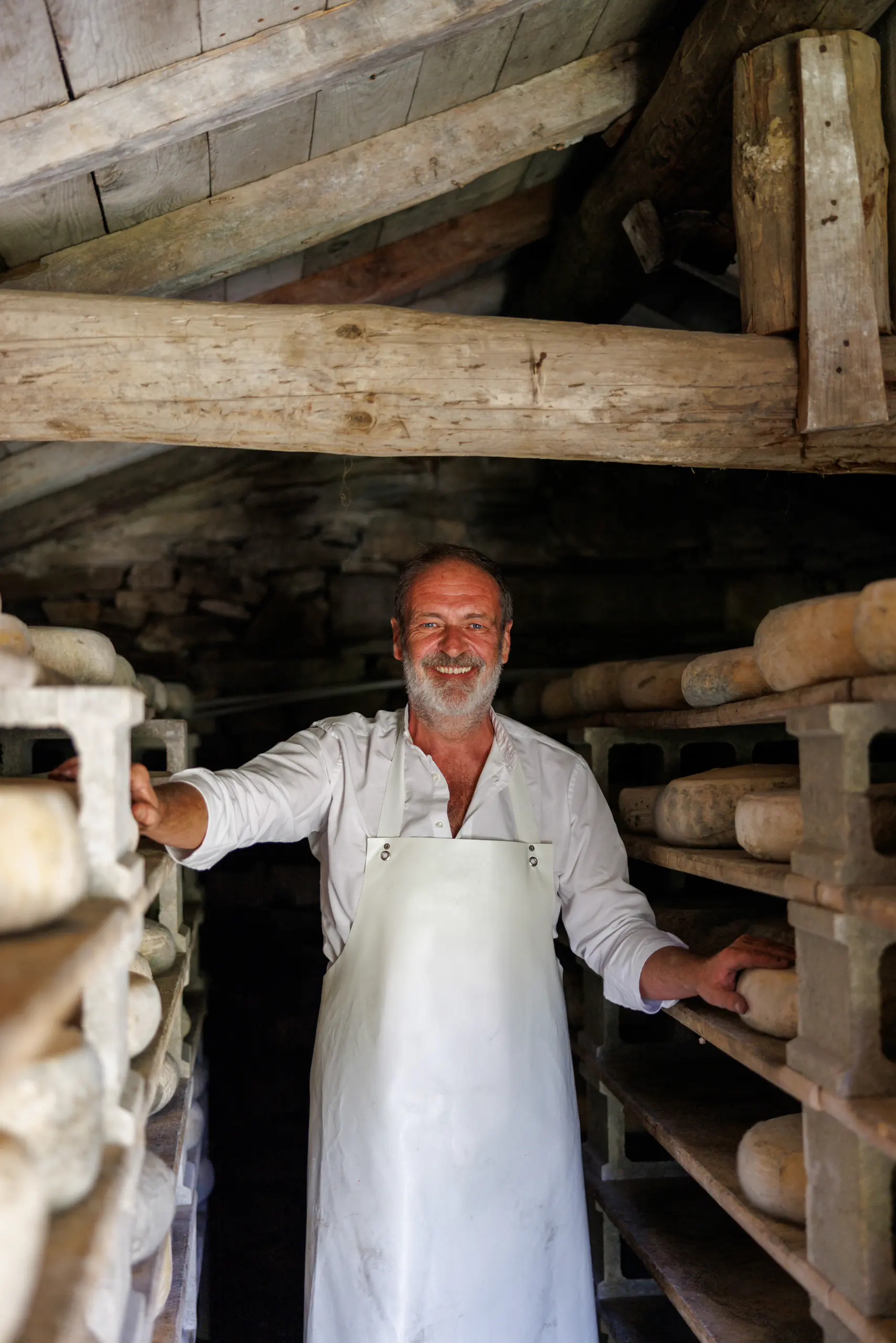 Older man in an apron among shelves of aging cheese, arms outstretched