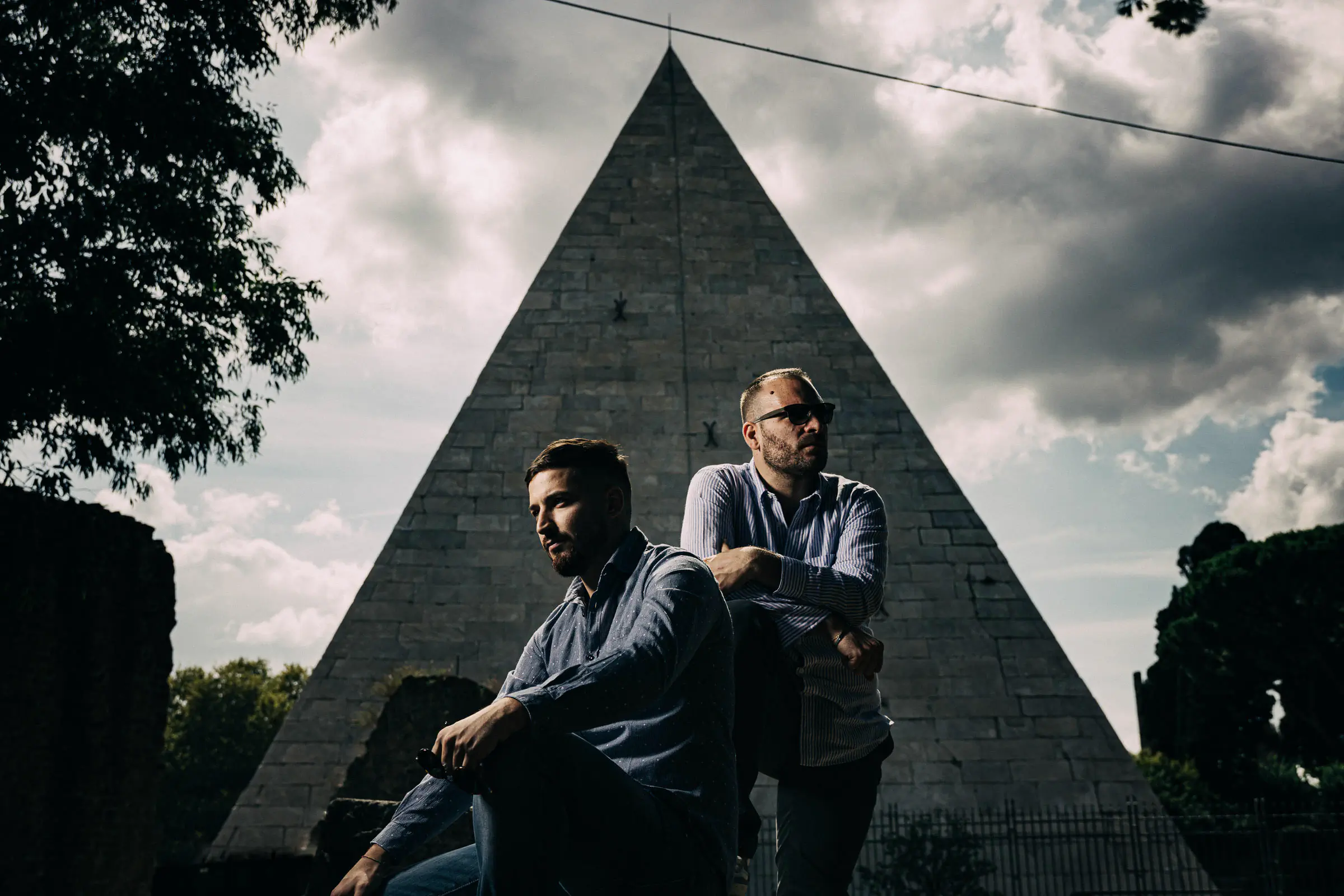 Two men seated at the base of a pyramid monument under a cloudy sky