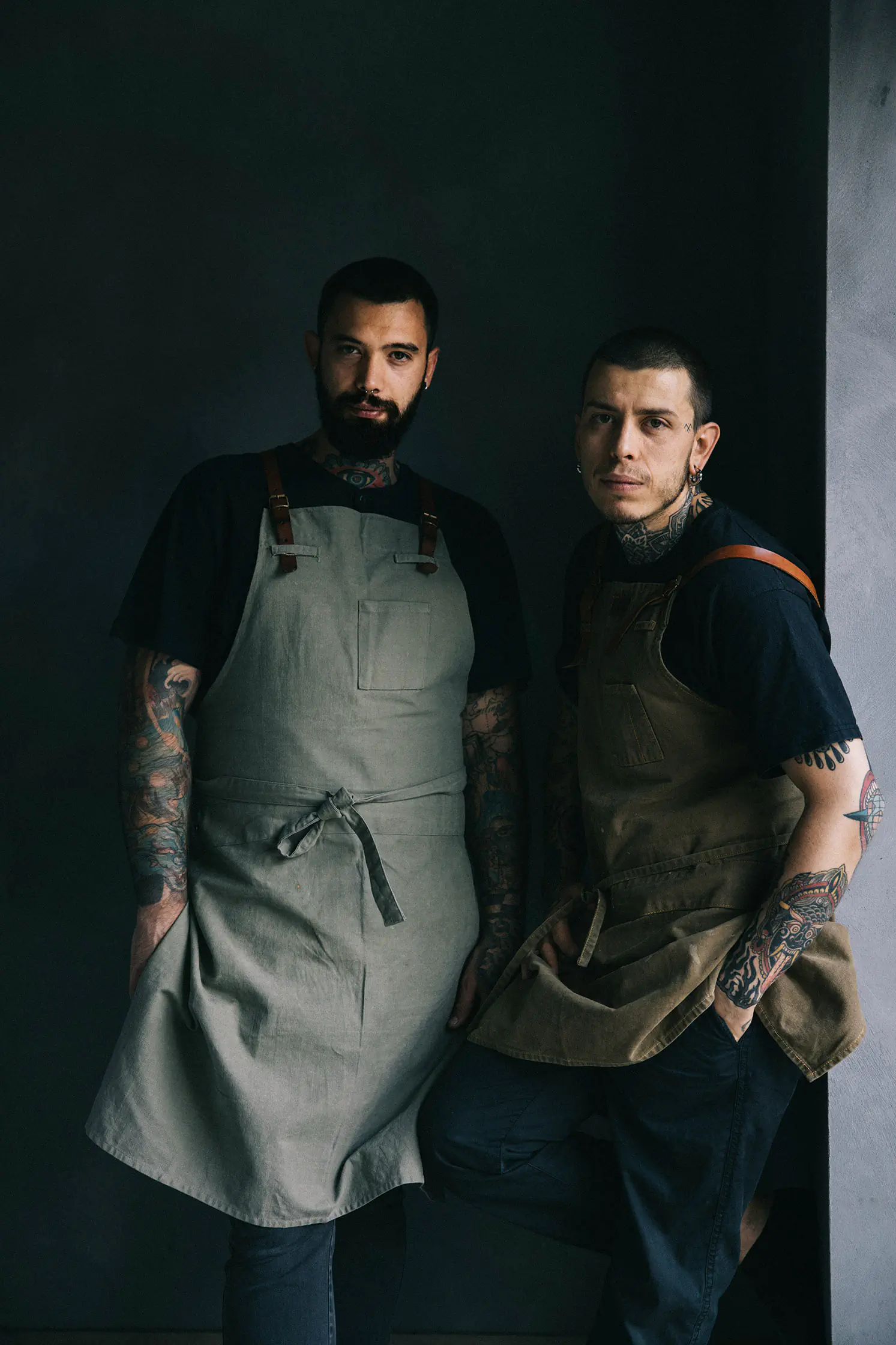 Two men in aprons posing against a dark wall