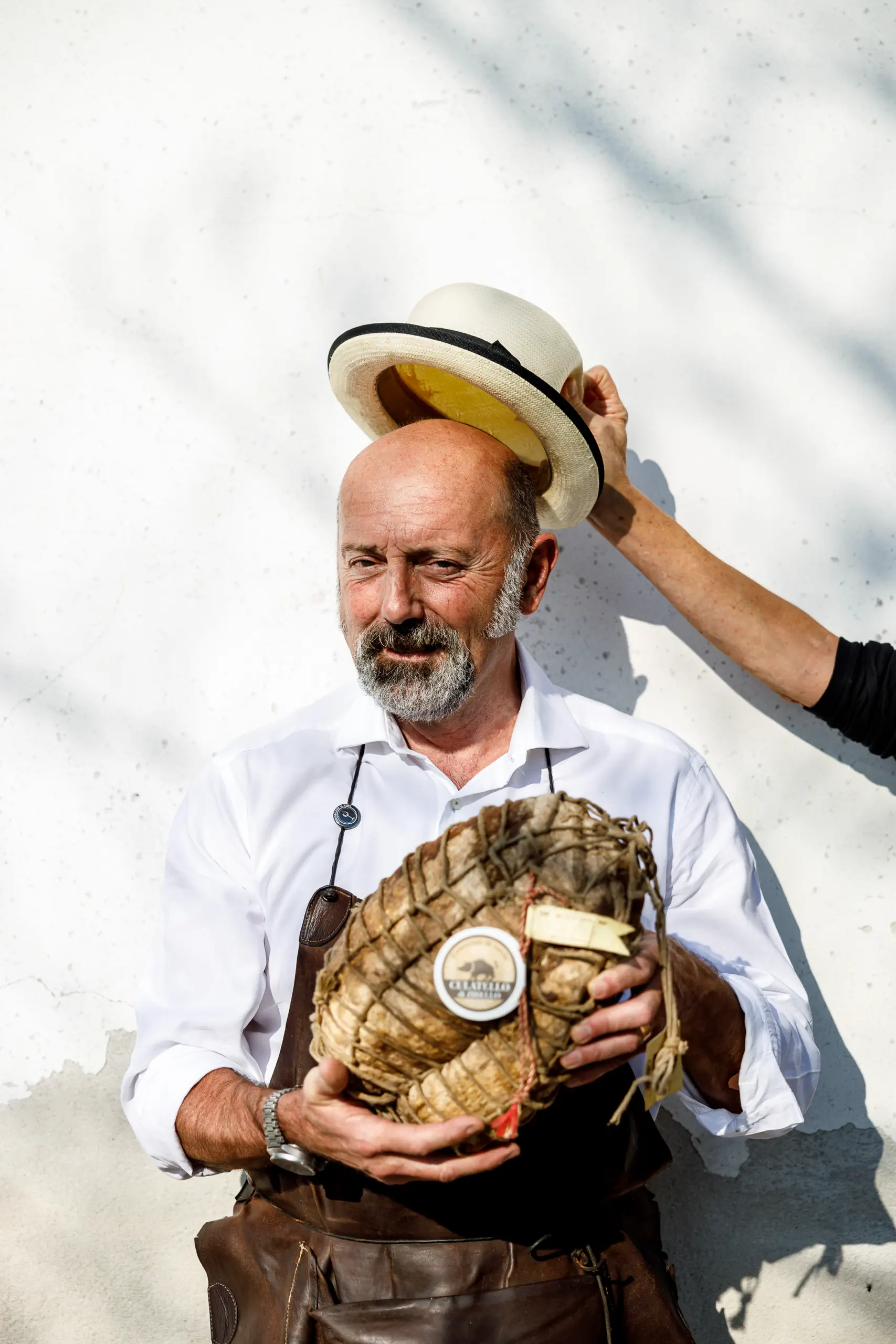 Man holding a wheel of cheese outdoors as a straw hat is placed on his head