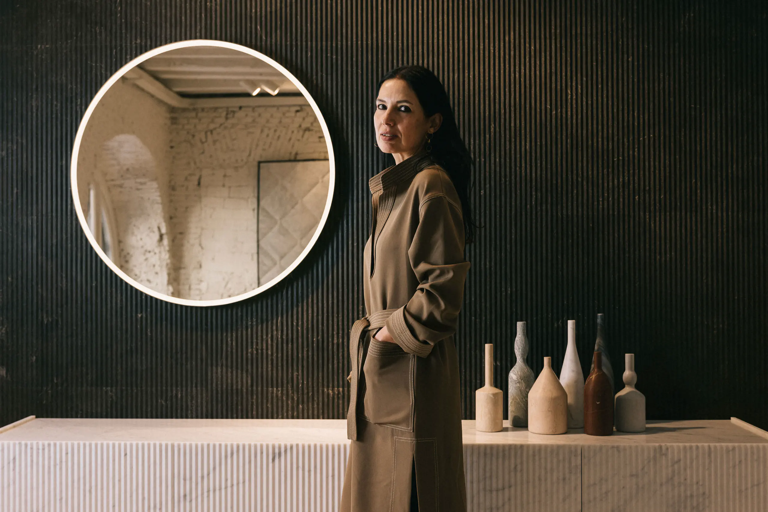Woman in a long coat by a round mirror and a console with sculptural vases