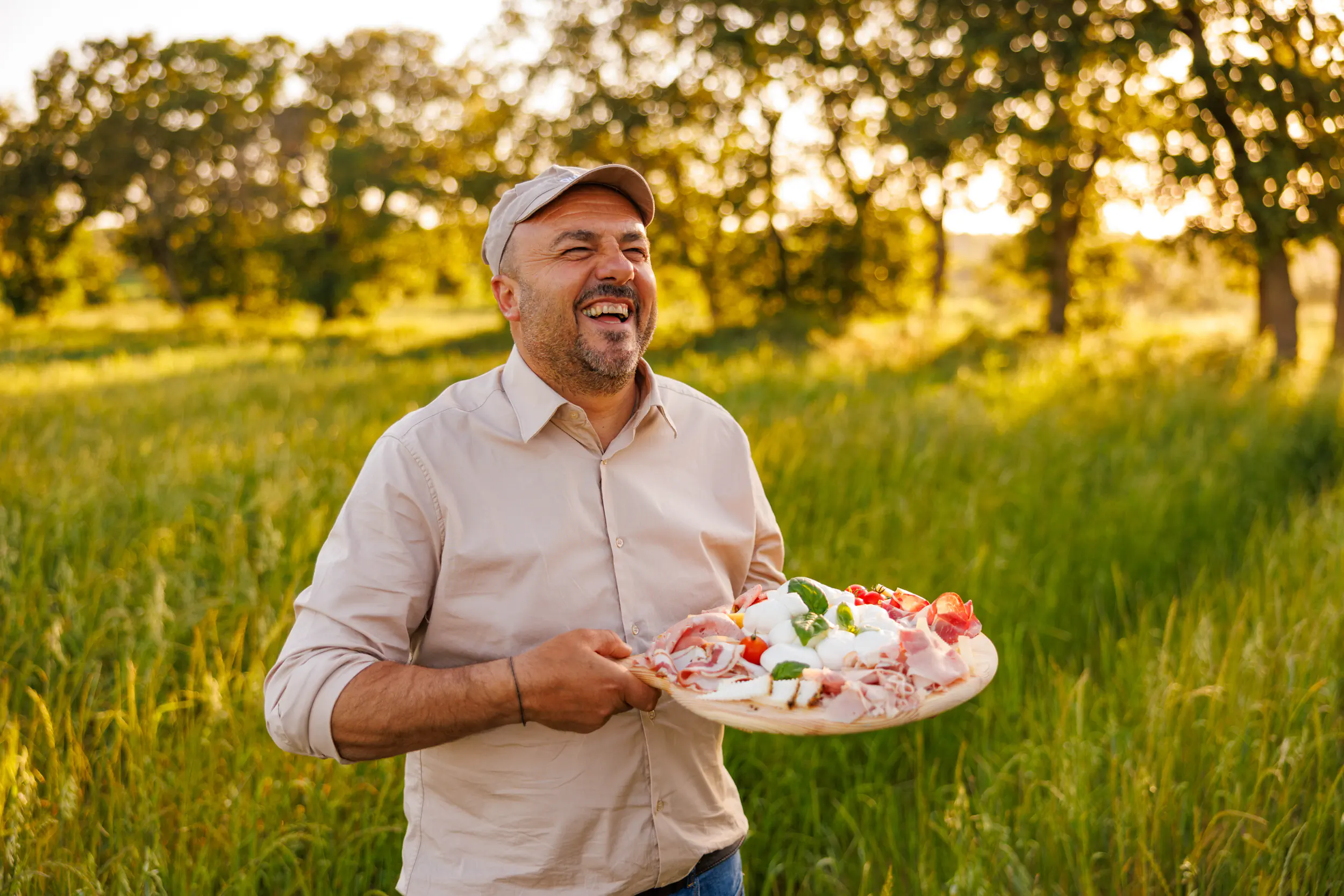 Smiling man in a cap holding a plate of food in a sunlit meadow