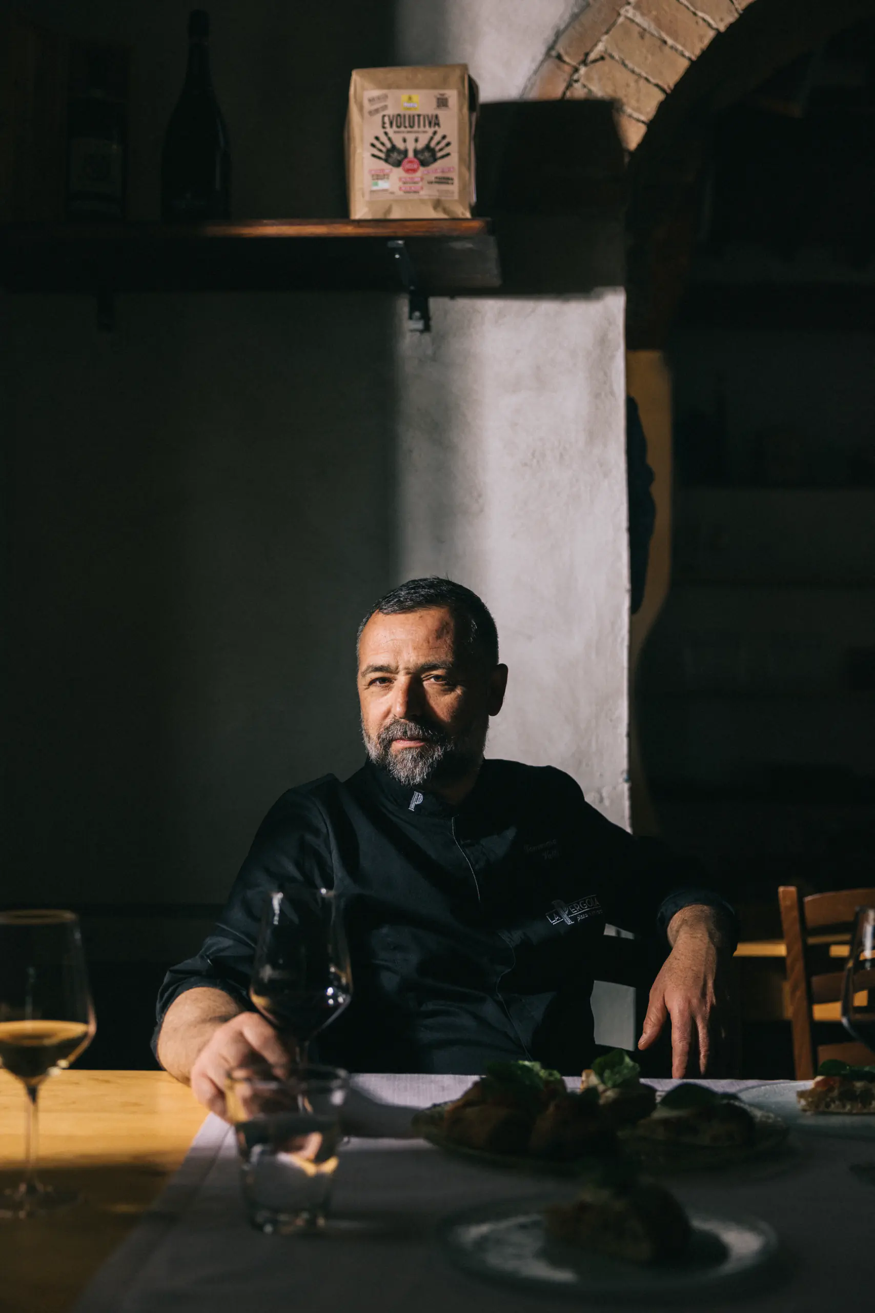 Man in black seated at a table with a glass of wine in low light