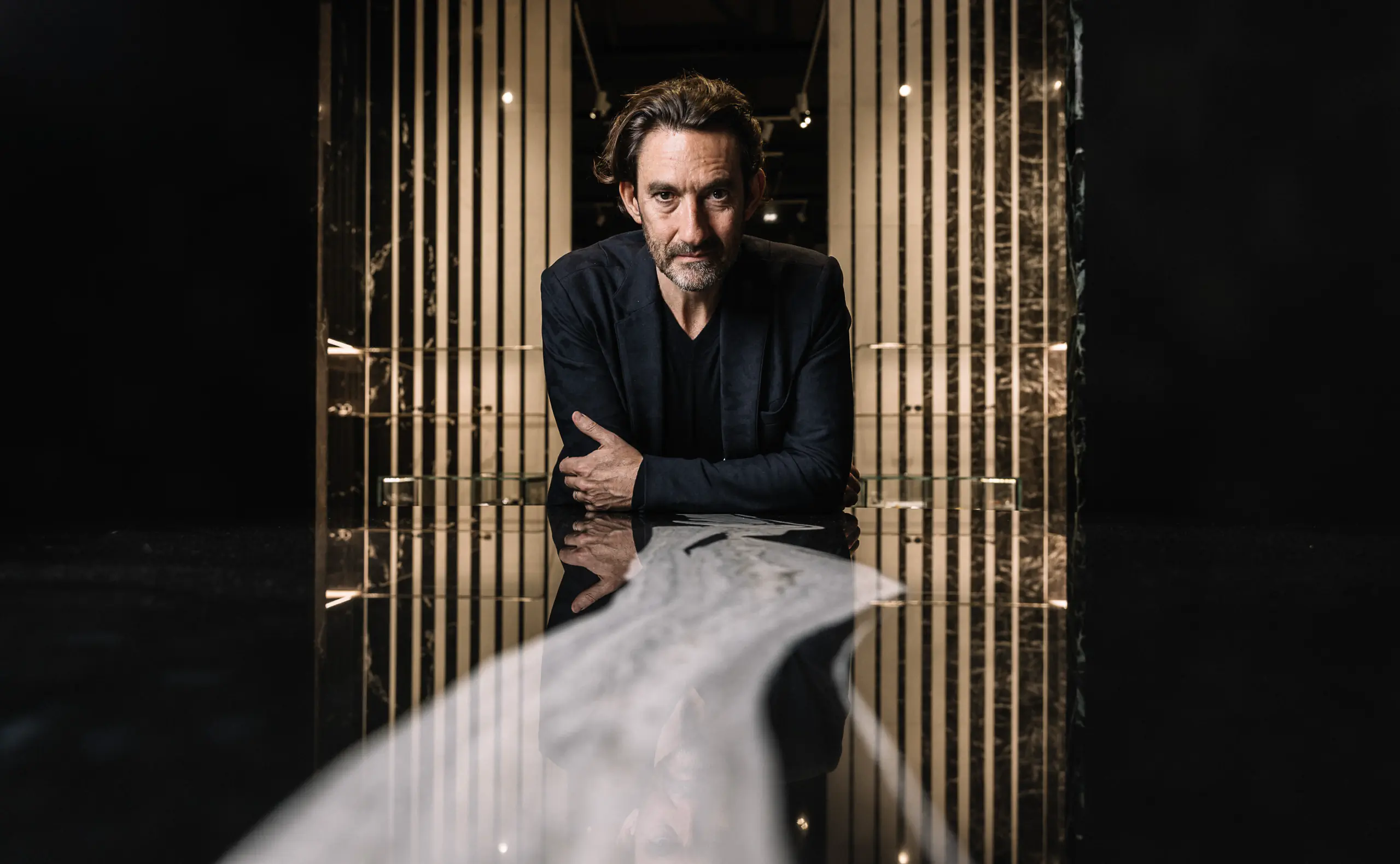 Man in black seated at a dark reflective table in a moody setting