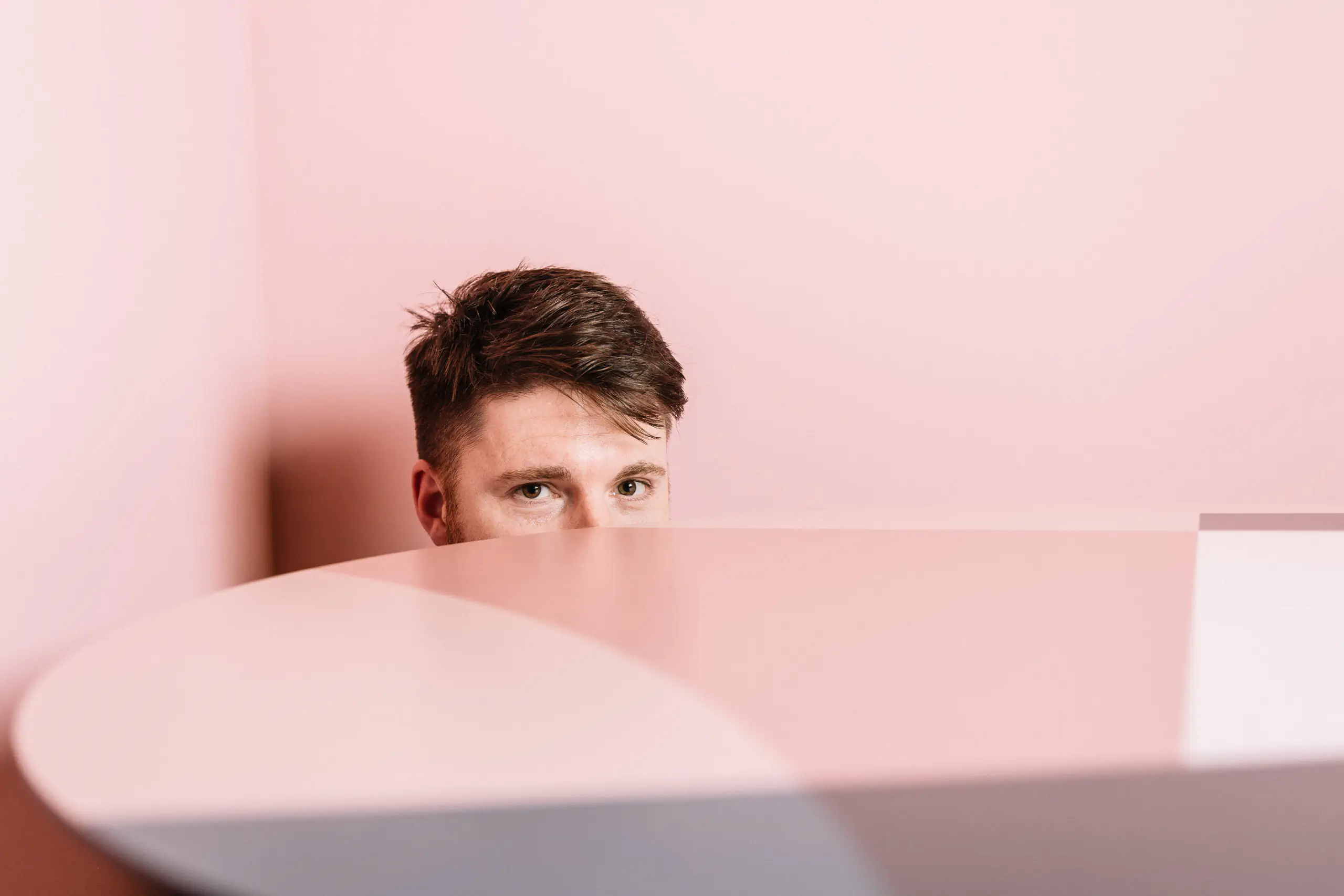 Young man peeking over a pink surface against a pink wall