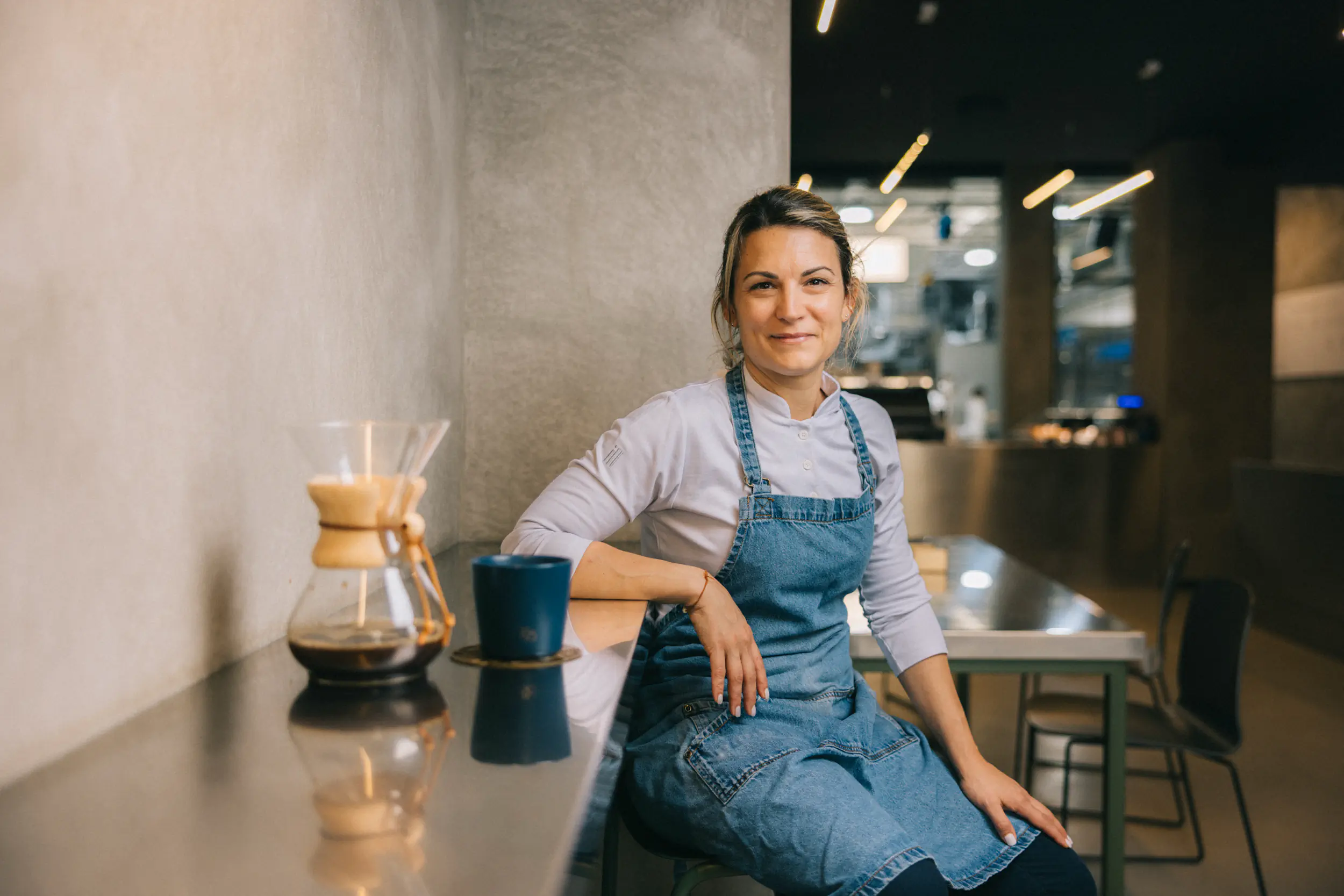 Woman in an apron seated at a café counter with a Chemex coffee maker
