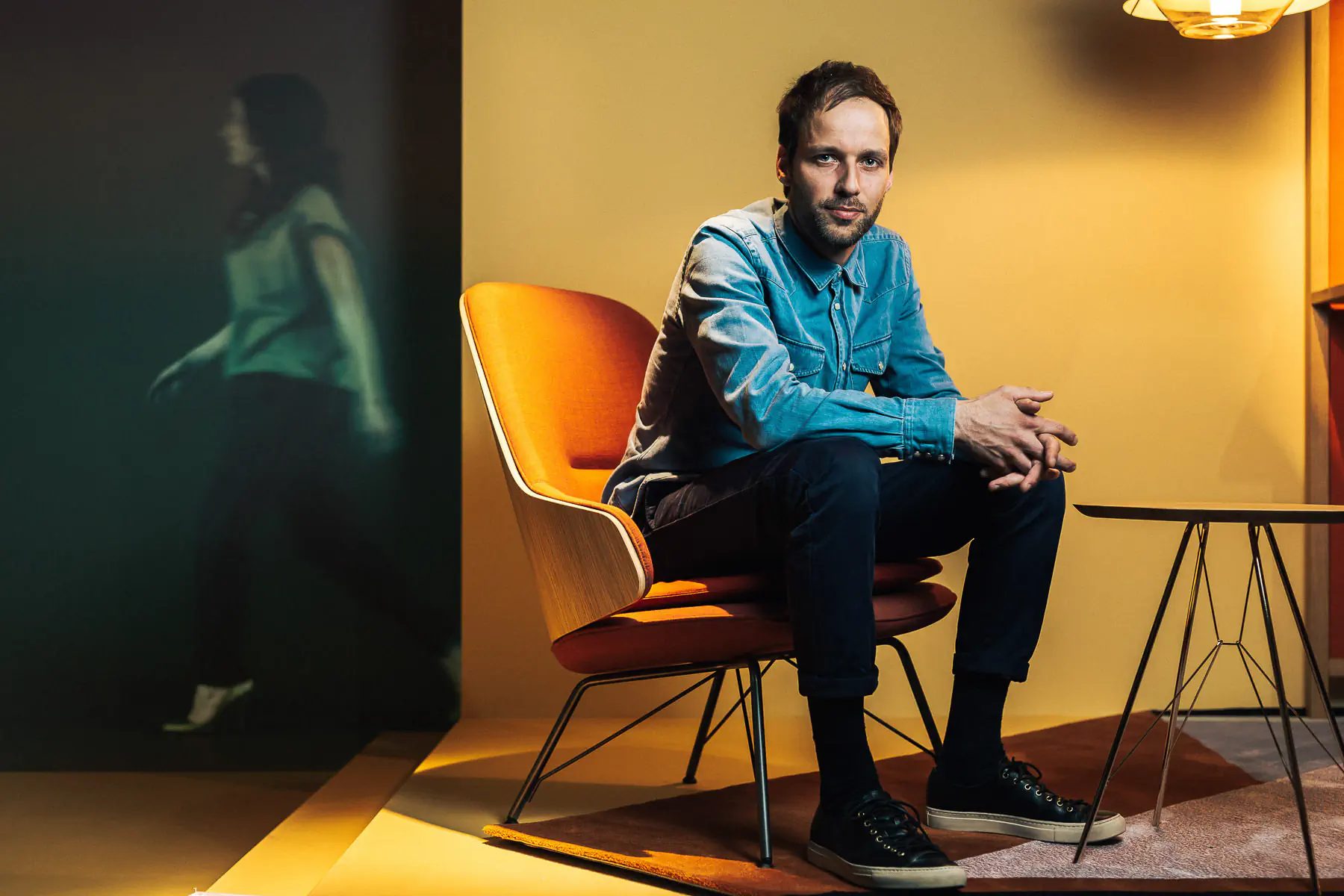 Man in a denim shirt seated on an orange chair in a warm-toned room