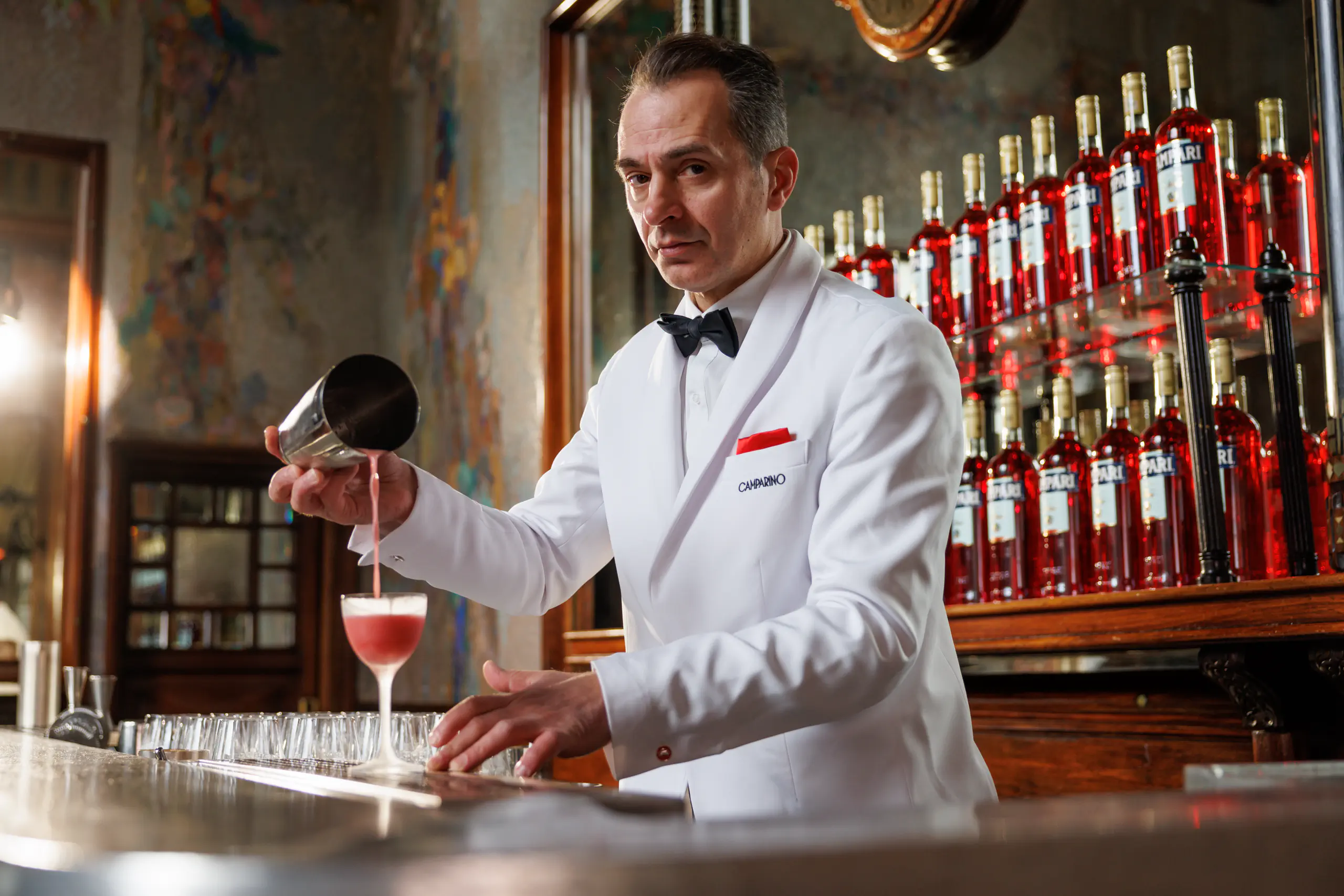 Bartender in a white tuxedo pouring a red cocktail behind a bar