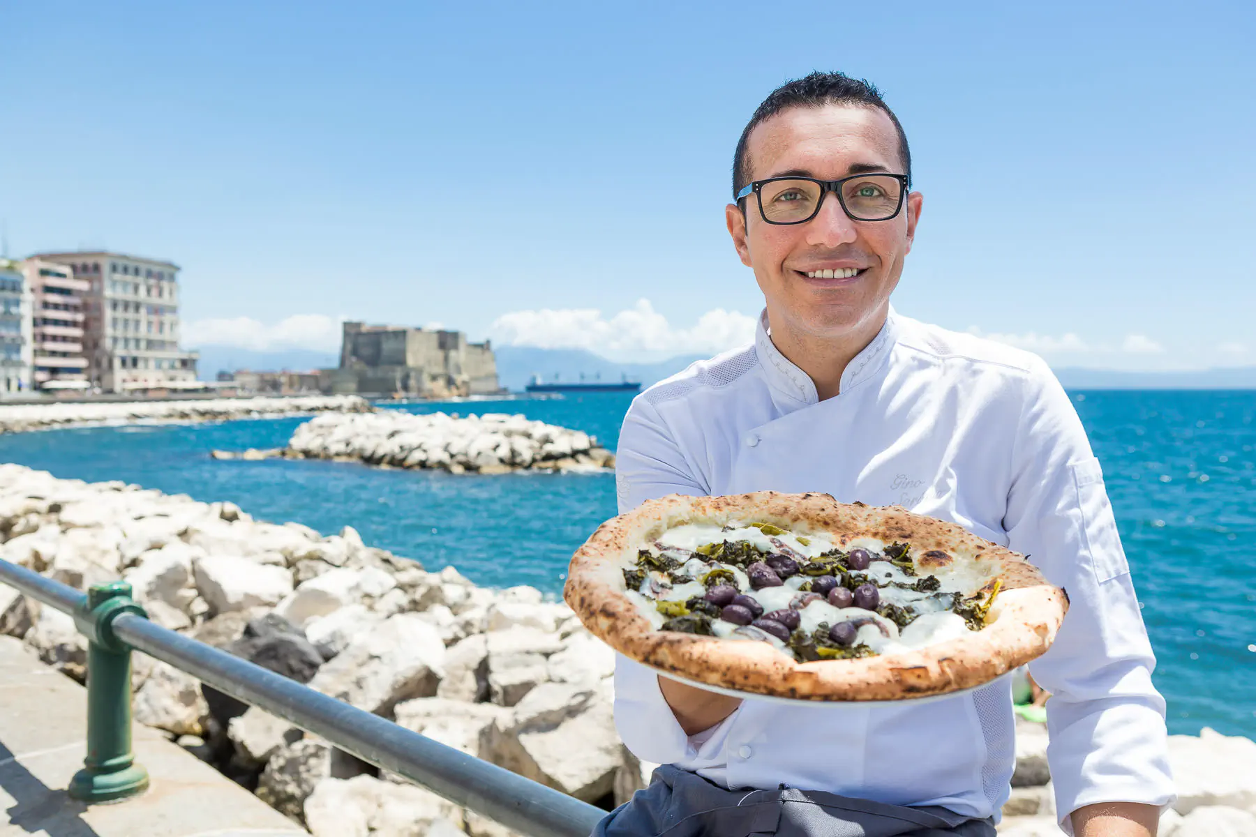 Smiling man holding a pizza on the Naples seafront with a castle behind