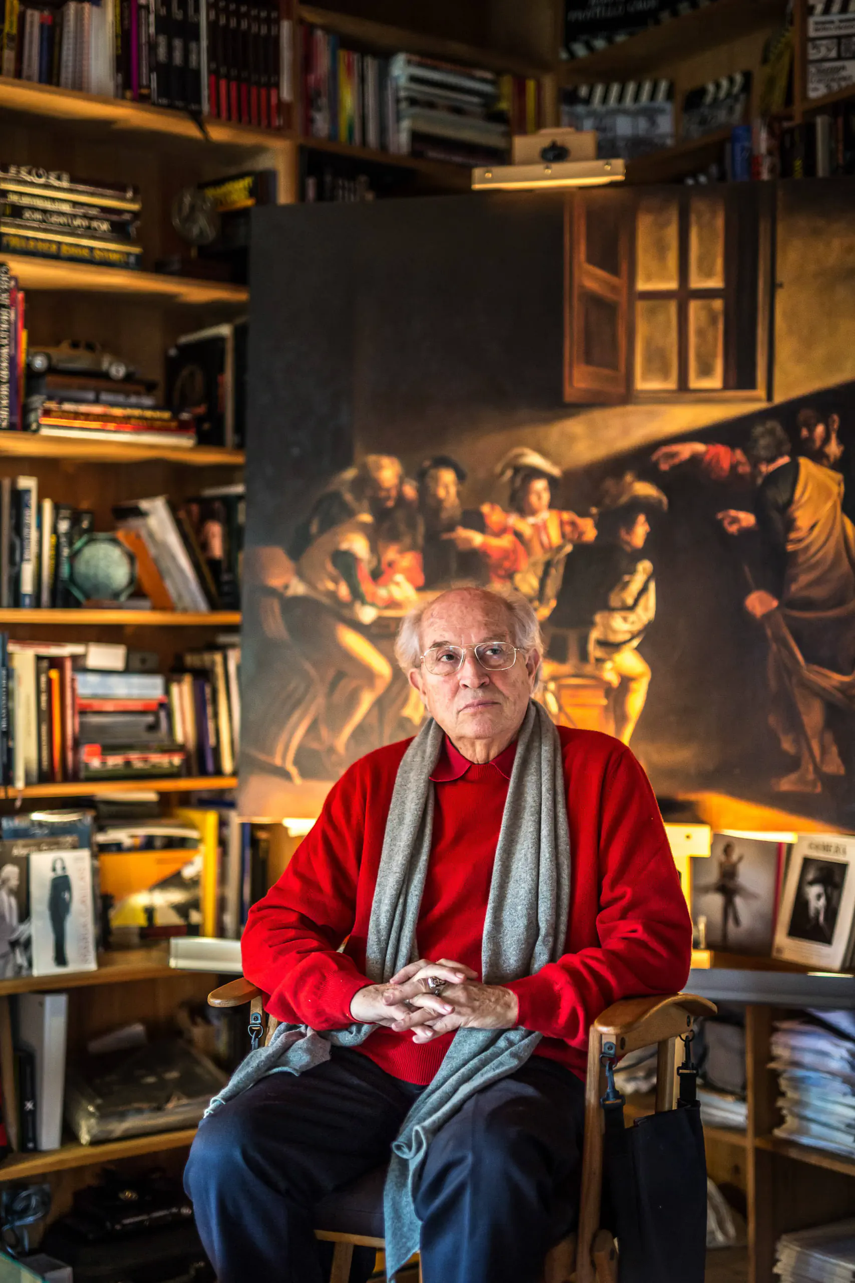 Older man in a red jacket seated in a book-lined study with a large painting